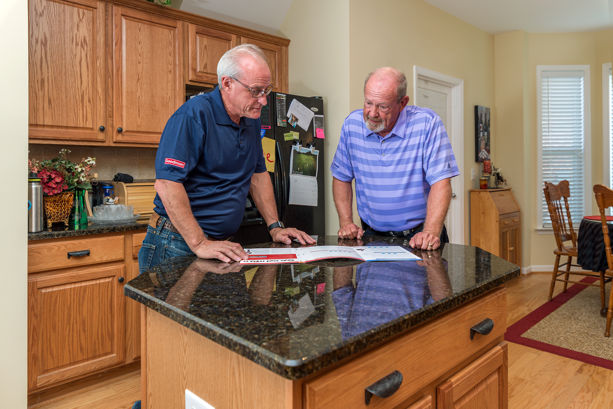Two older men review documents or a calendar at a kitchen island in a bright, modern kitchen with wooden cabinets and a black granite countertop.
