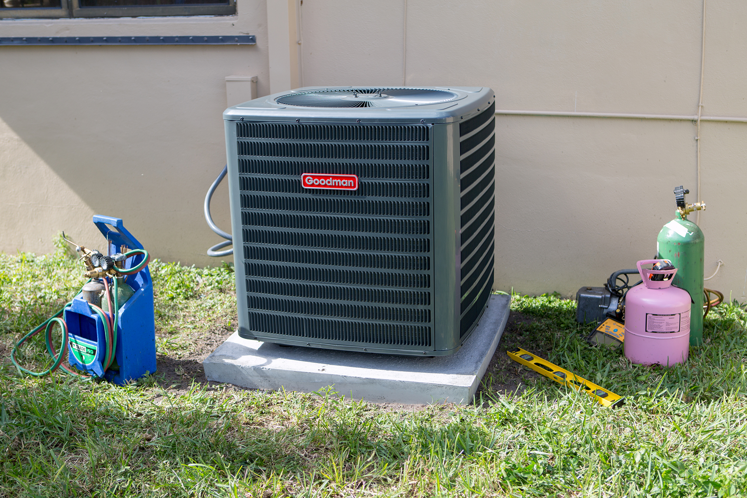 An outdoor air conditioning unit with a Goodman logo, surrounded by tools and refrigerant tanks on grass and concrete.