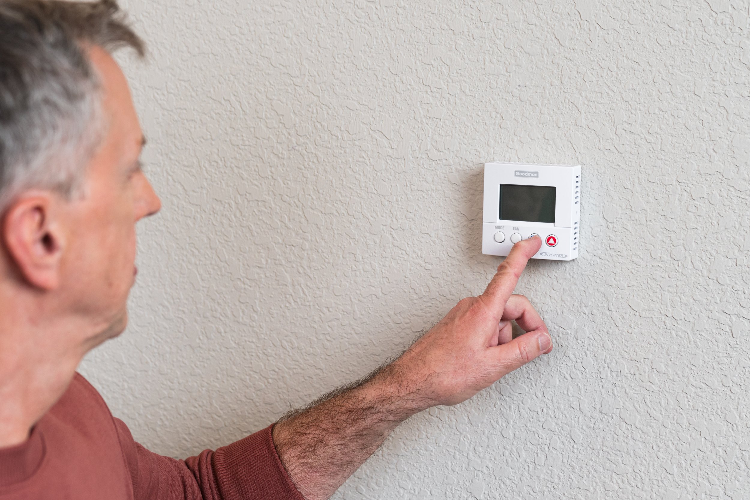 A man in a brown shirt adjusts a white thermostat on a textured beige wall.
