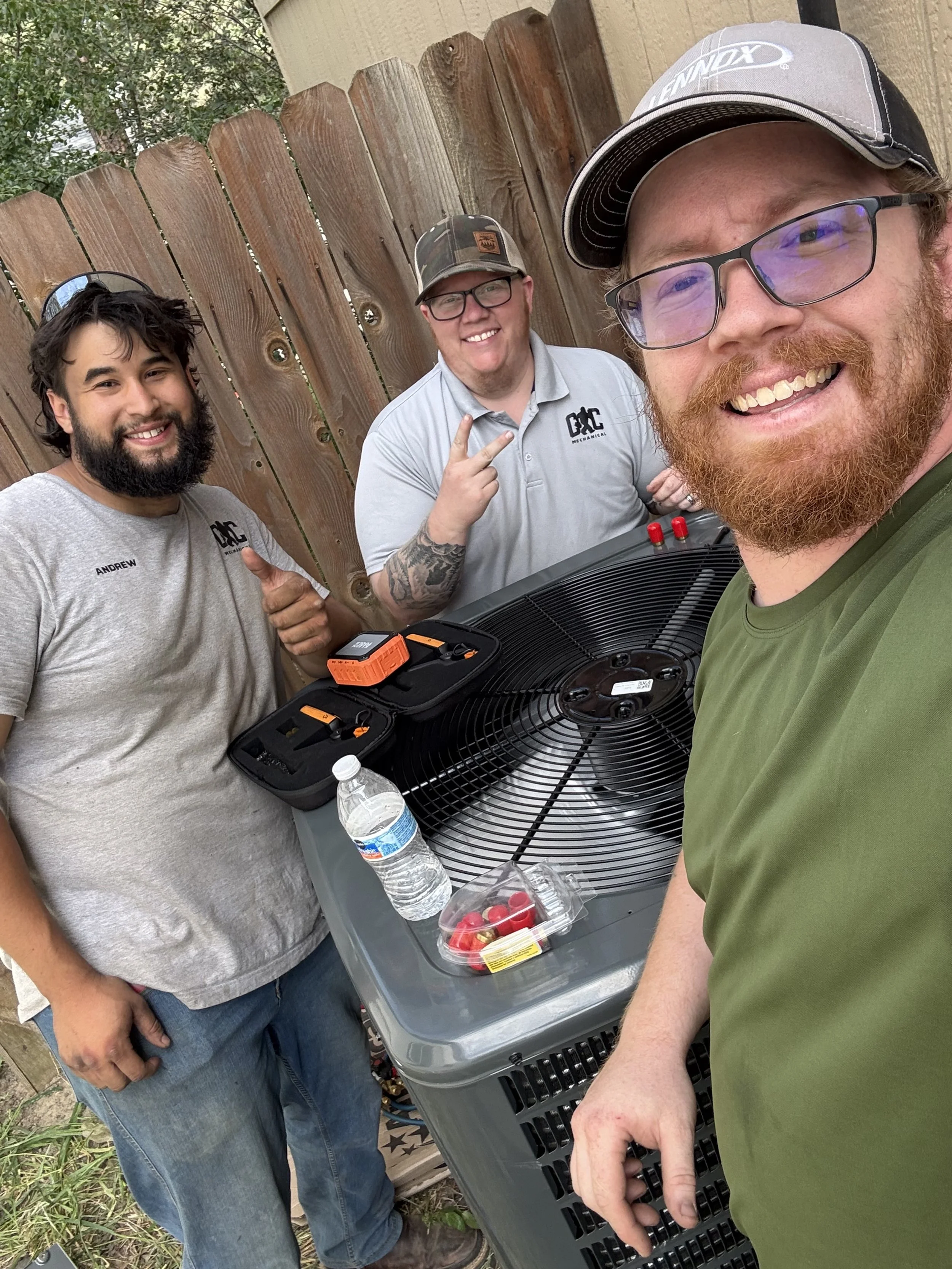 Three men smiling outdoors near a wooden fence, taking a selfie. They are standing around an HVAC unit with tools..