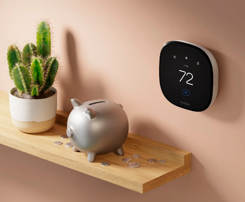 A wooden shelf with a potted cactus, a piggy bank, and scattered coins, with a digital thermostat on the wall displaying 72 degrees.