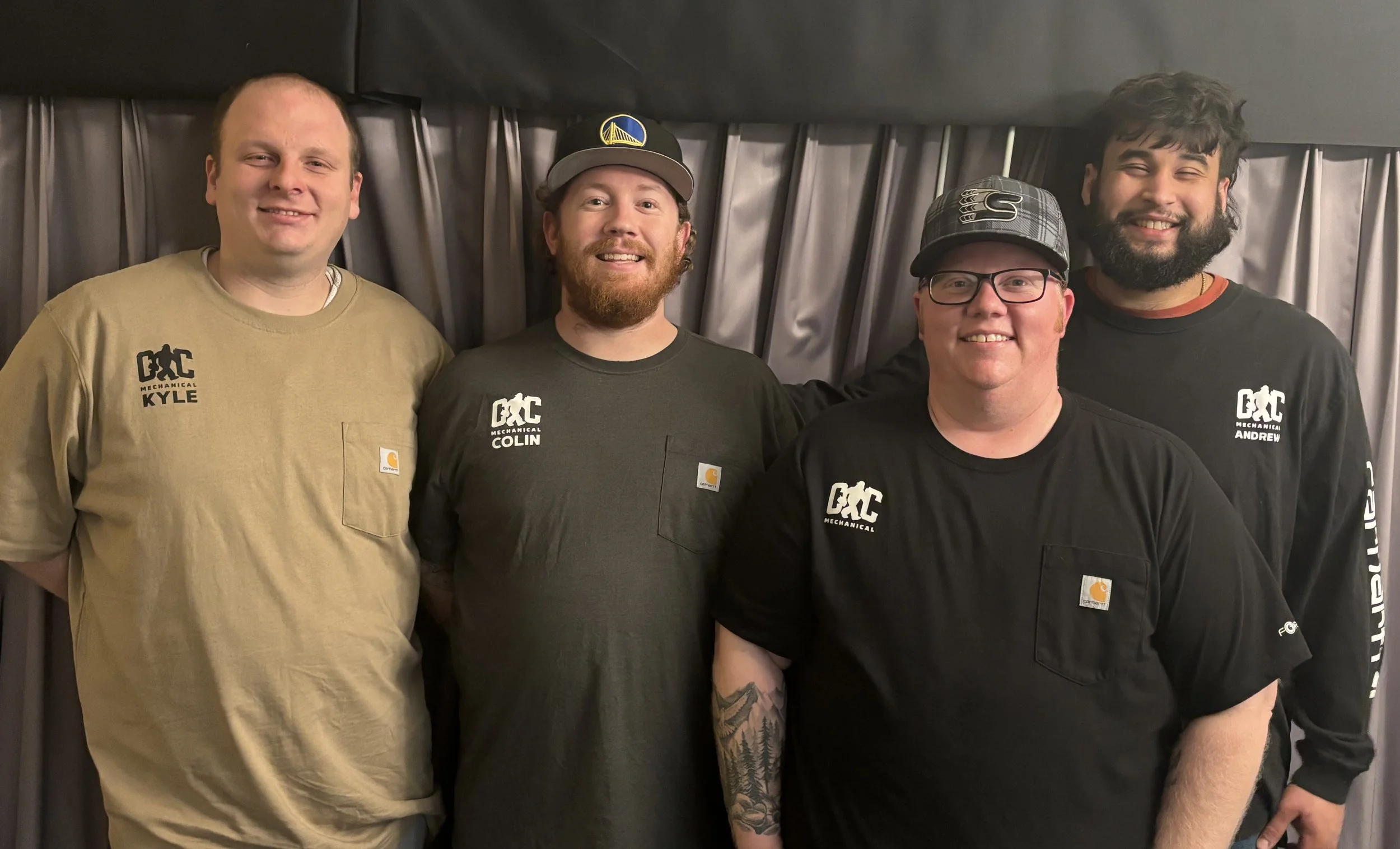 Four men standing together indoors with a dark curtain in the background, all smiling at the camera. They are wearing casual work shirts with logos and name tags.