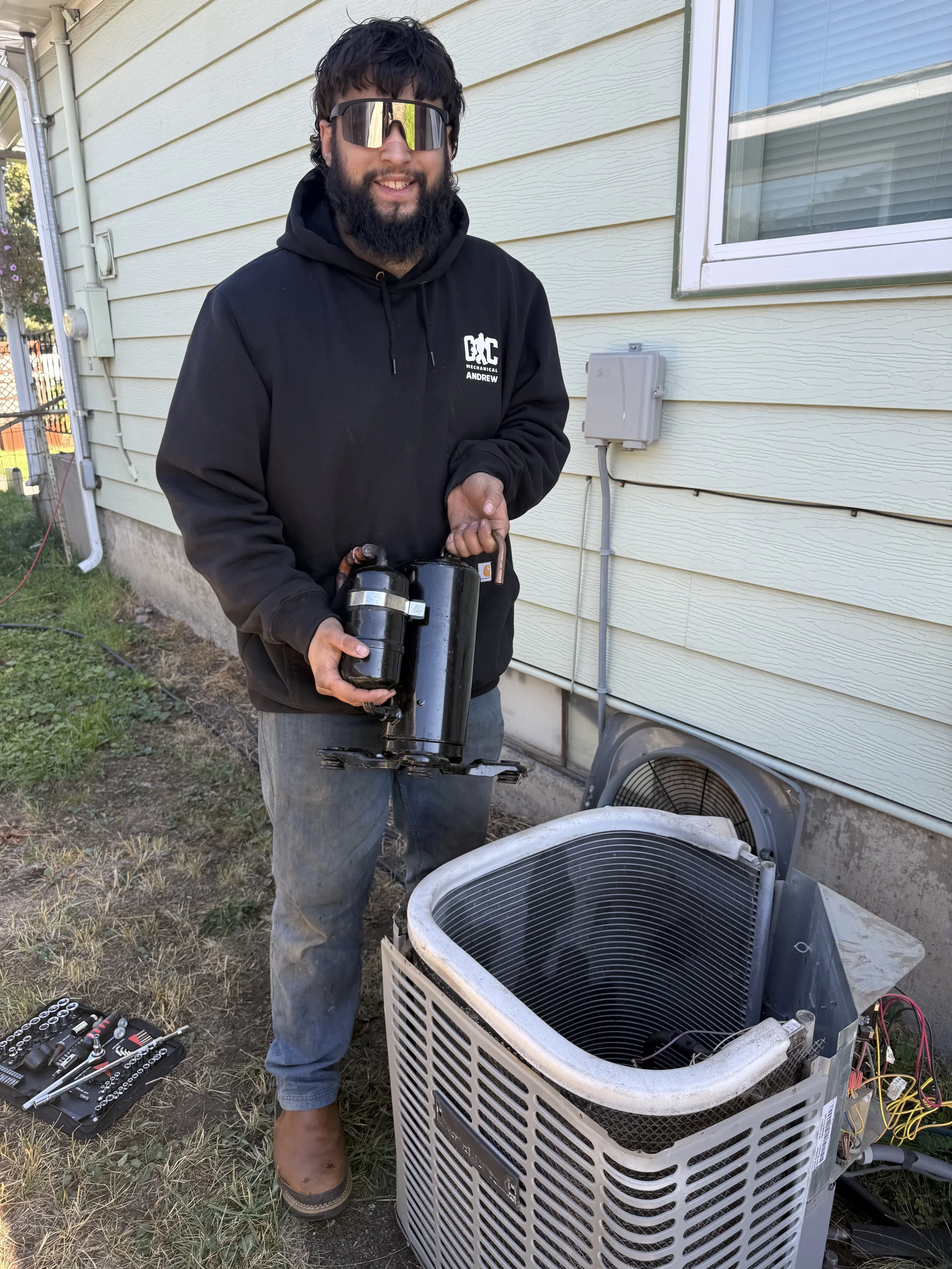 A man with sunglasses and a beard wearing a black hoodie labeled 'C Mechanical Andrew' is holding a refrigerant recovery machine. He is standing outside next to an open air conditioning unit on the ground, with tools nearby, in front of a beige house with horizontal siding.