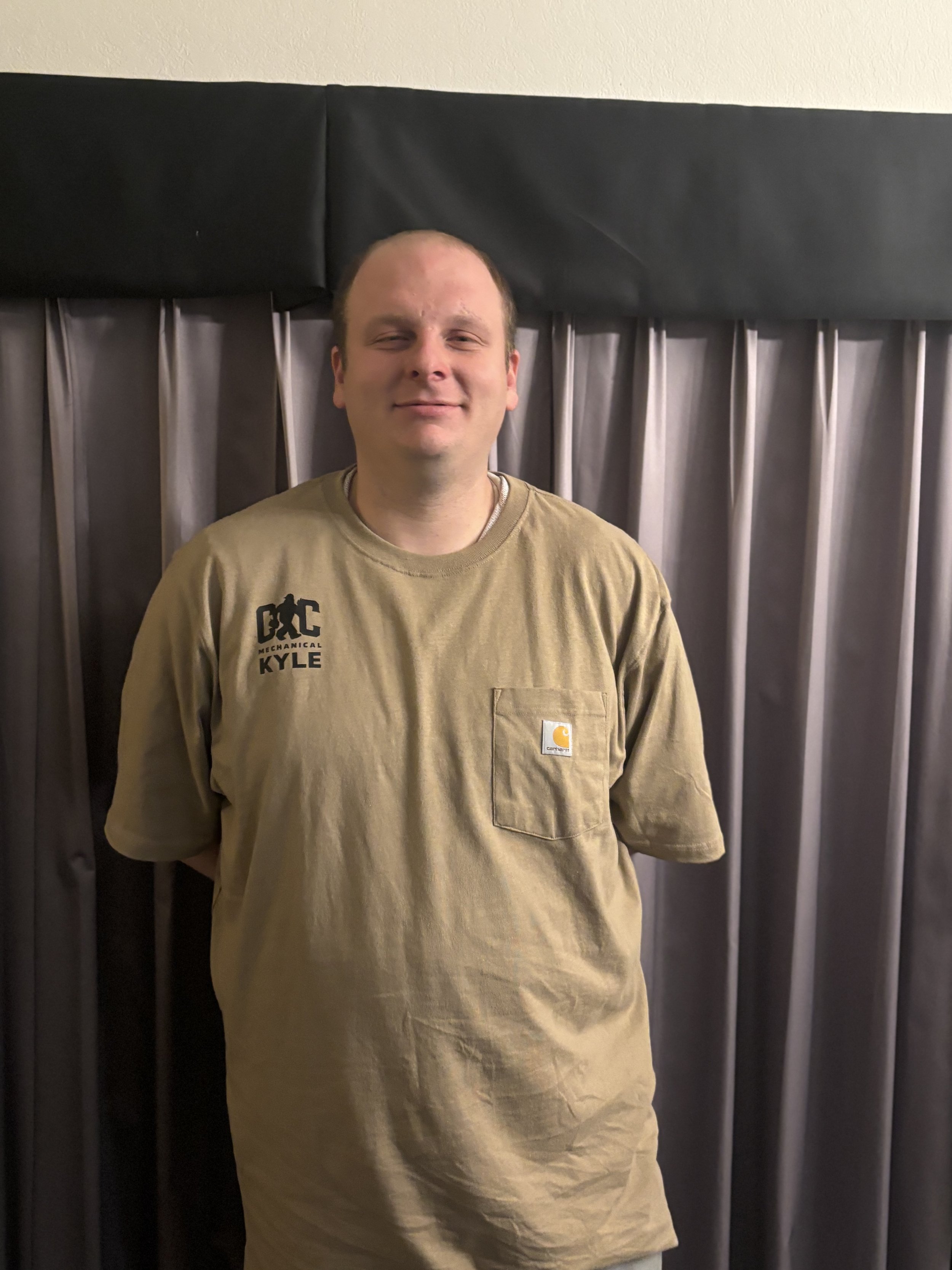 A young man with light skin, short hair, and a slight smile, standing in front of a dark curtain backdrop. He is wearing a tan Carhartt T-shirt with black text on the left side that reads 'D C MECHANICAL KYLE,' and a small pocket on the right side with a Carhartt logo.