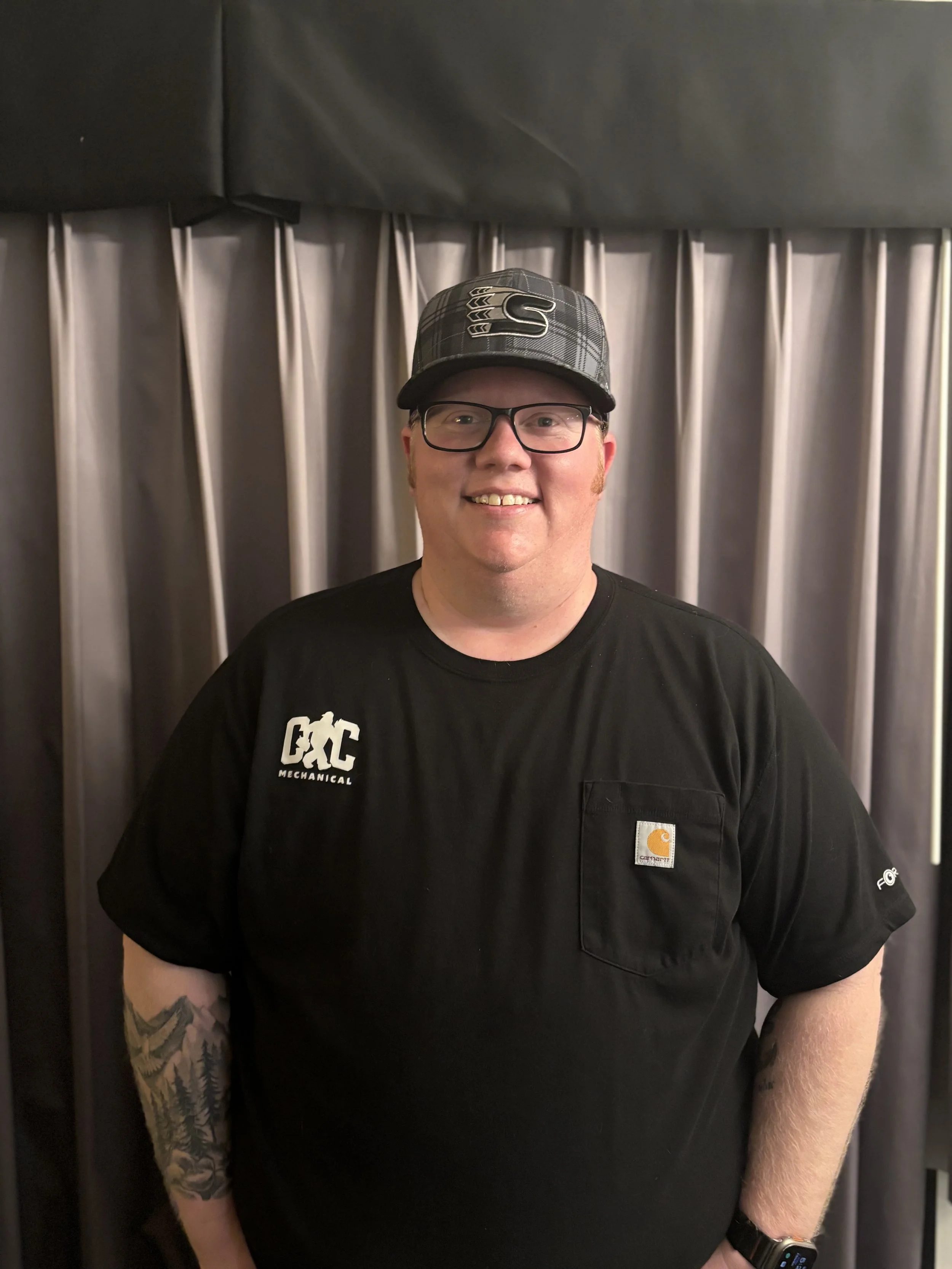 A smiling man wearing glasses, a black Carhartt t-shirt, a black cap with a Spokane Chiefs logo, and a watch, standing in front of a curtain.