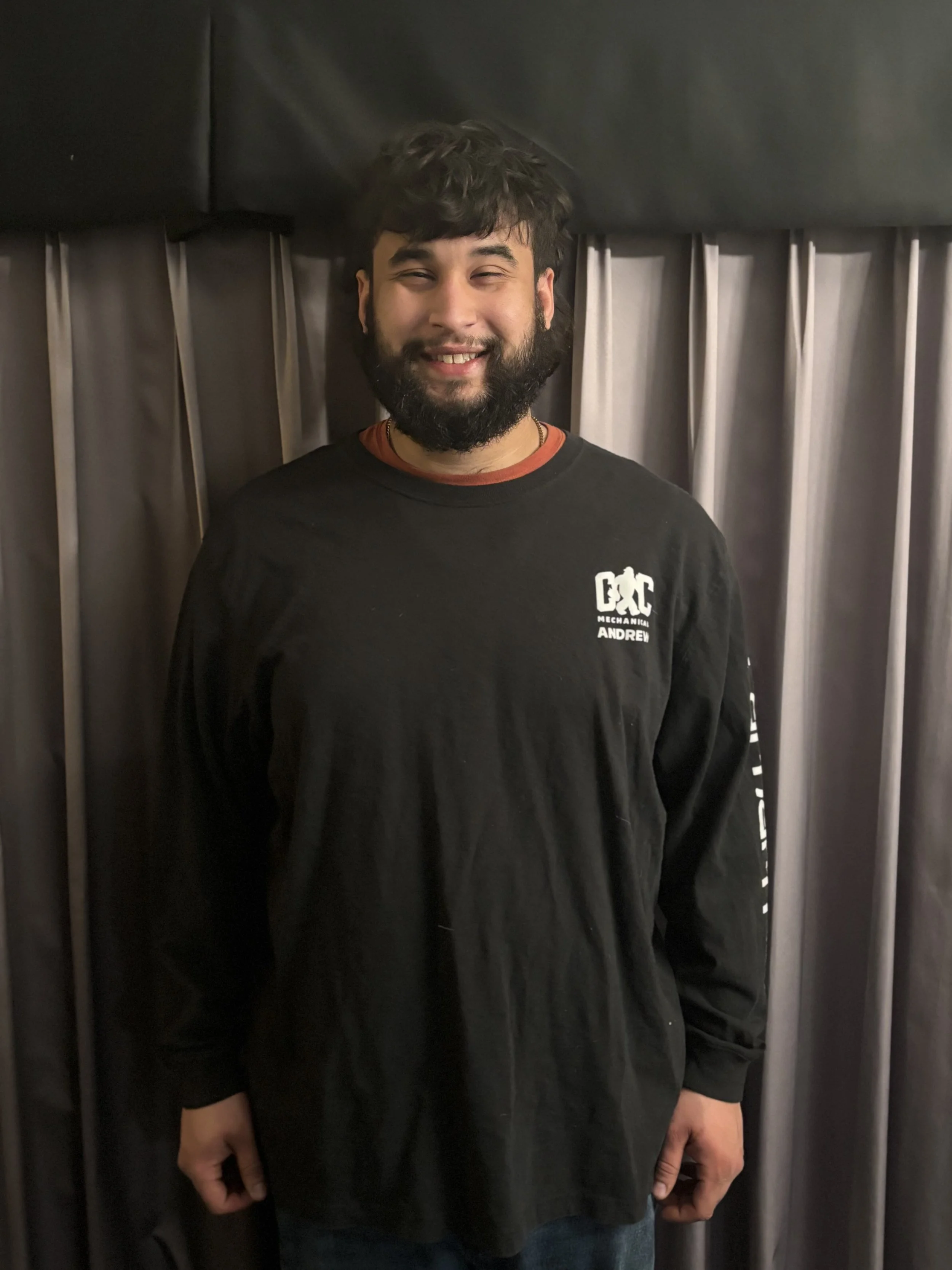 A young man with dark hair and a beard, smiling, standing in front of a gray curtain, wearing a black long-sleeved shirt with white text.