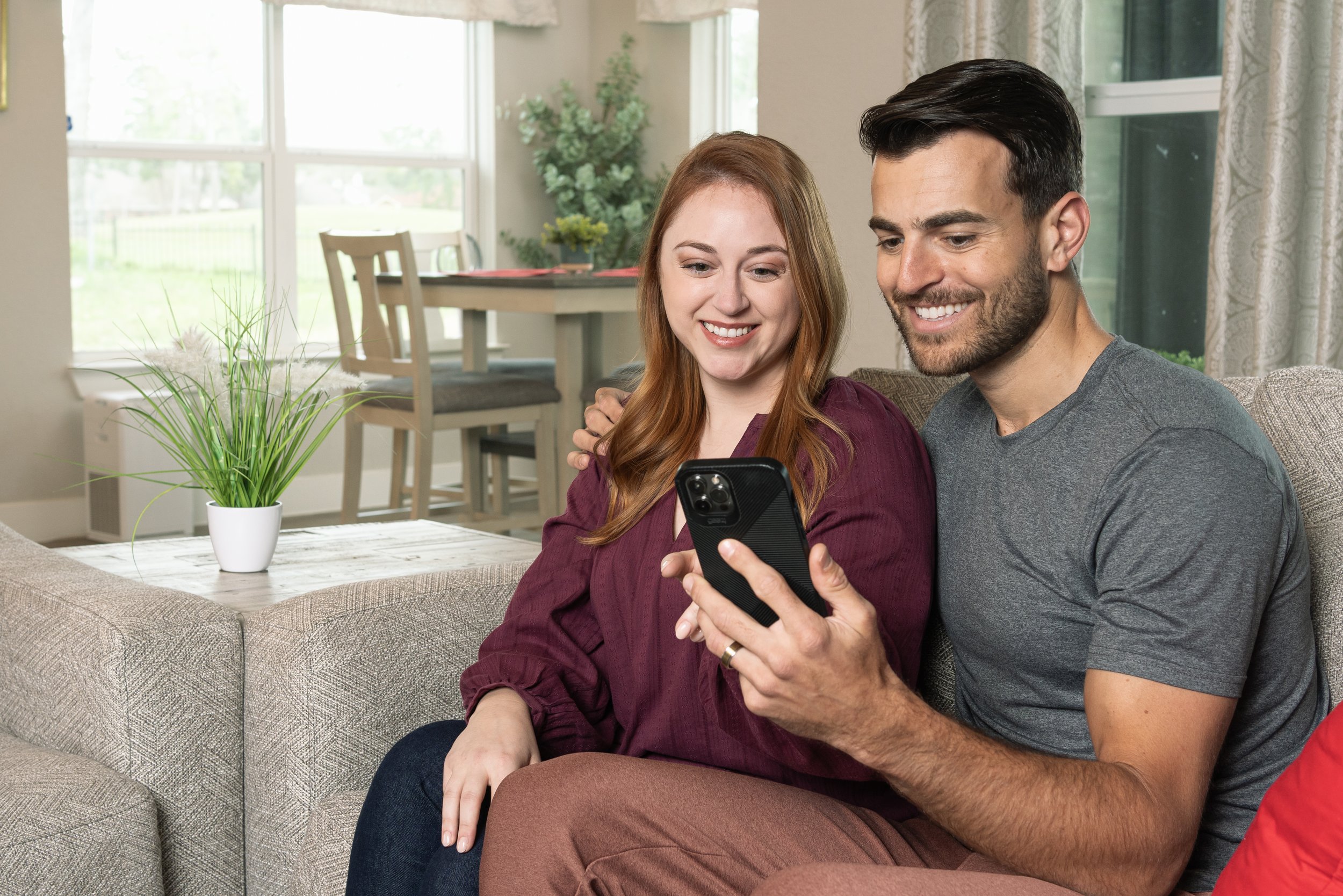 A smiling man and woman sitting on a beige couch, looking at a smartphone together in a well-lit living room with a window and a dining area in the background.