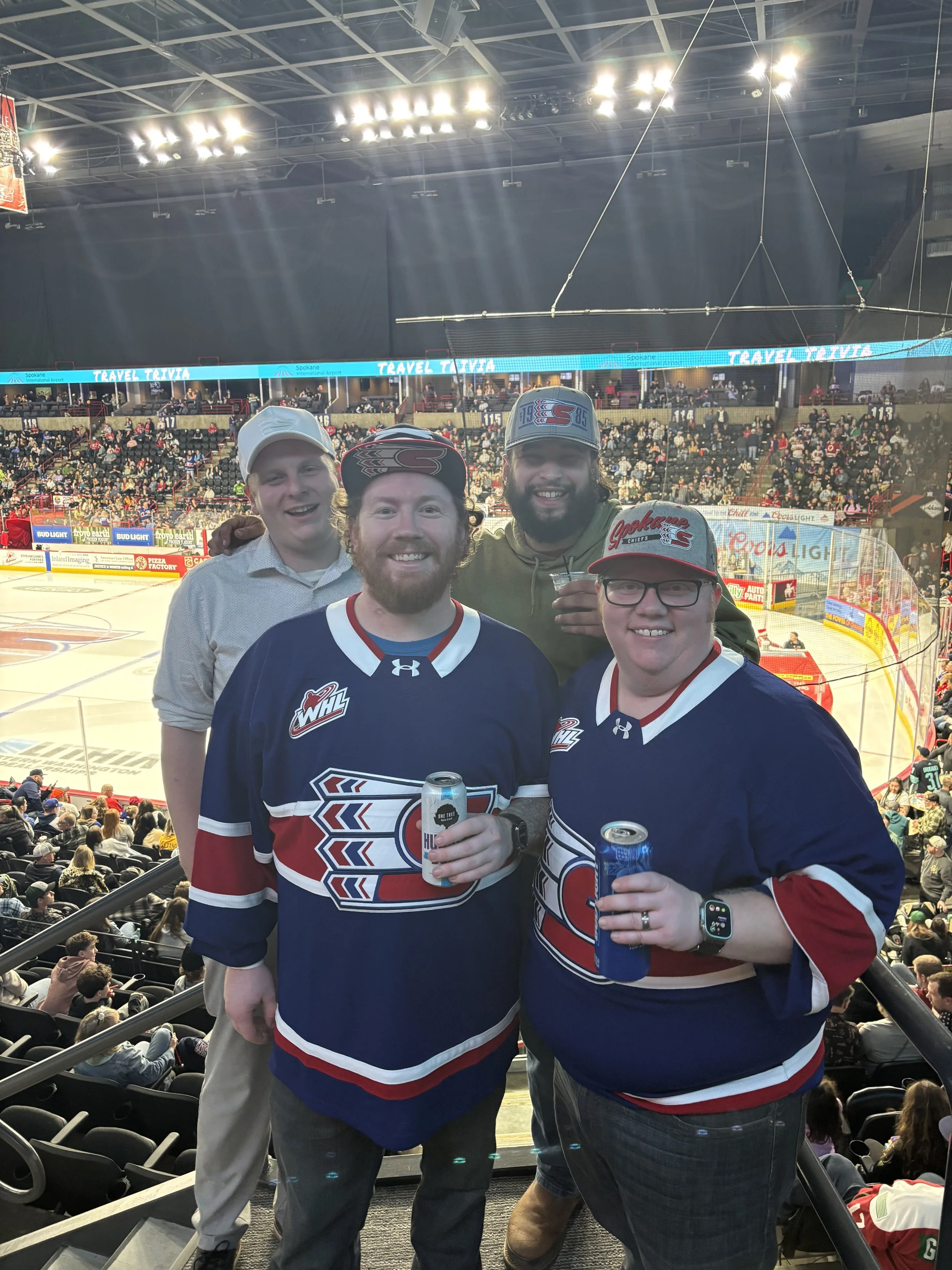 Four hockey fans posing for a photo inside an arena with an ice rink and audience in the background, three wearing hockey jerseys and hats, two holding drinks.