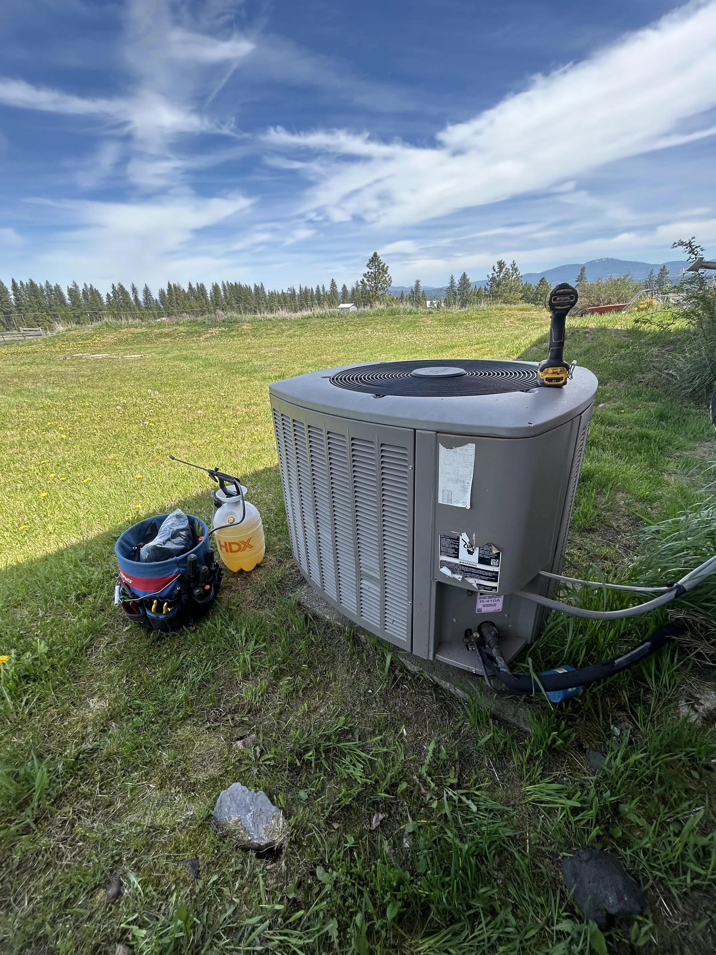 An outdoor air conditioning unit on a grassy area with a tool bag and a yellow HDX sprayer nearby, under a partly cloudy sky with mountains in the distance.