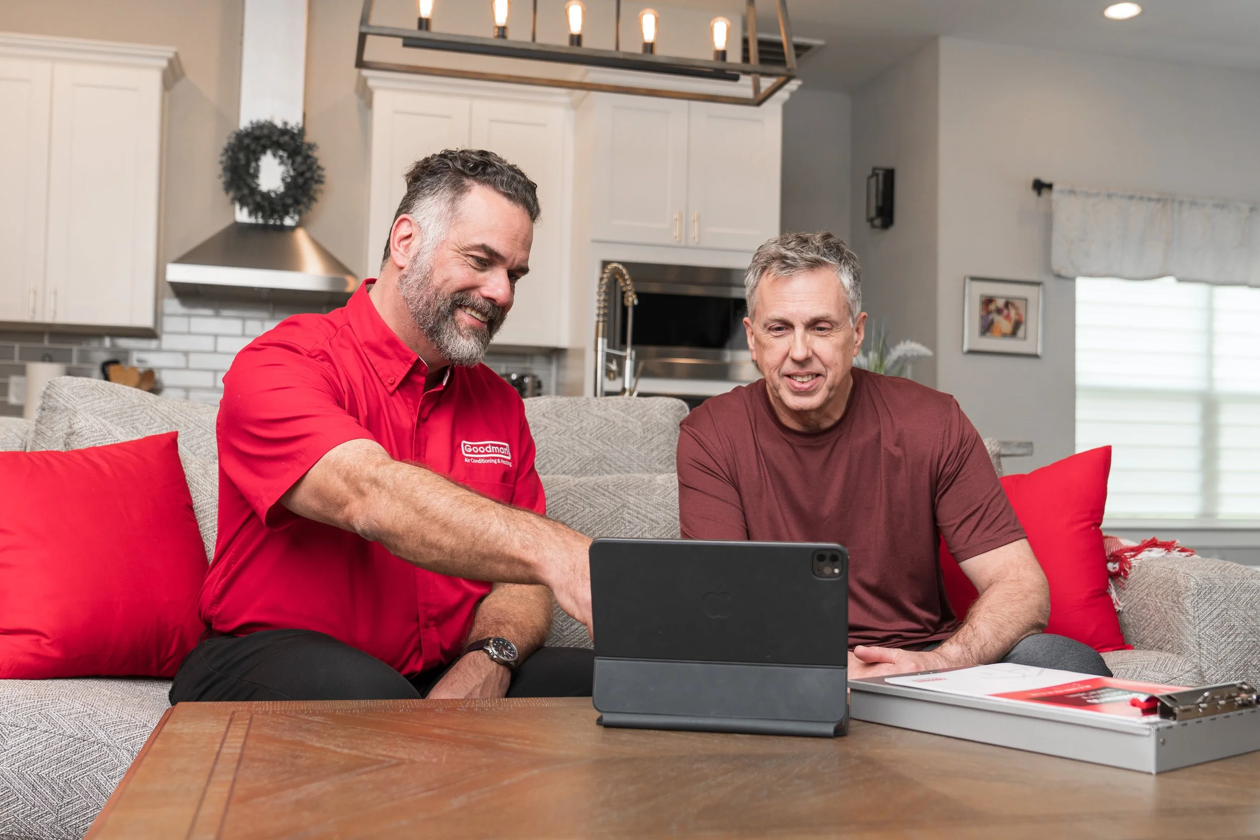 Two men sitting on a beige couch looking at a tablet on a coffee table, with a red pillow behind them in a bright living room.
