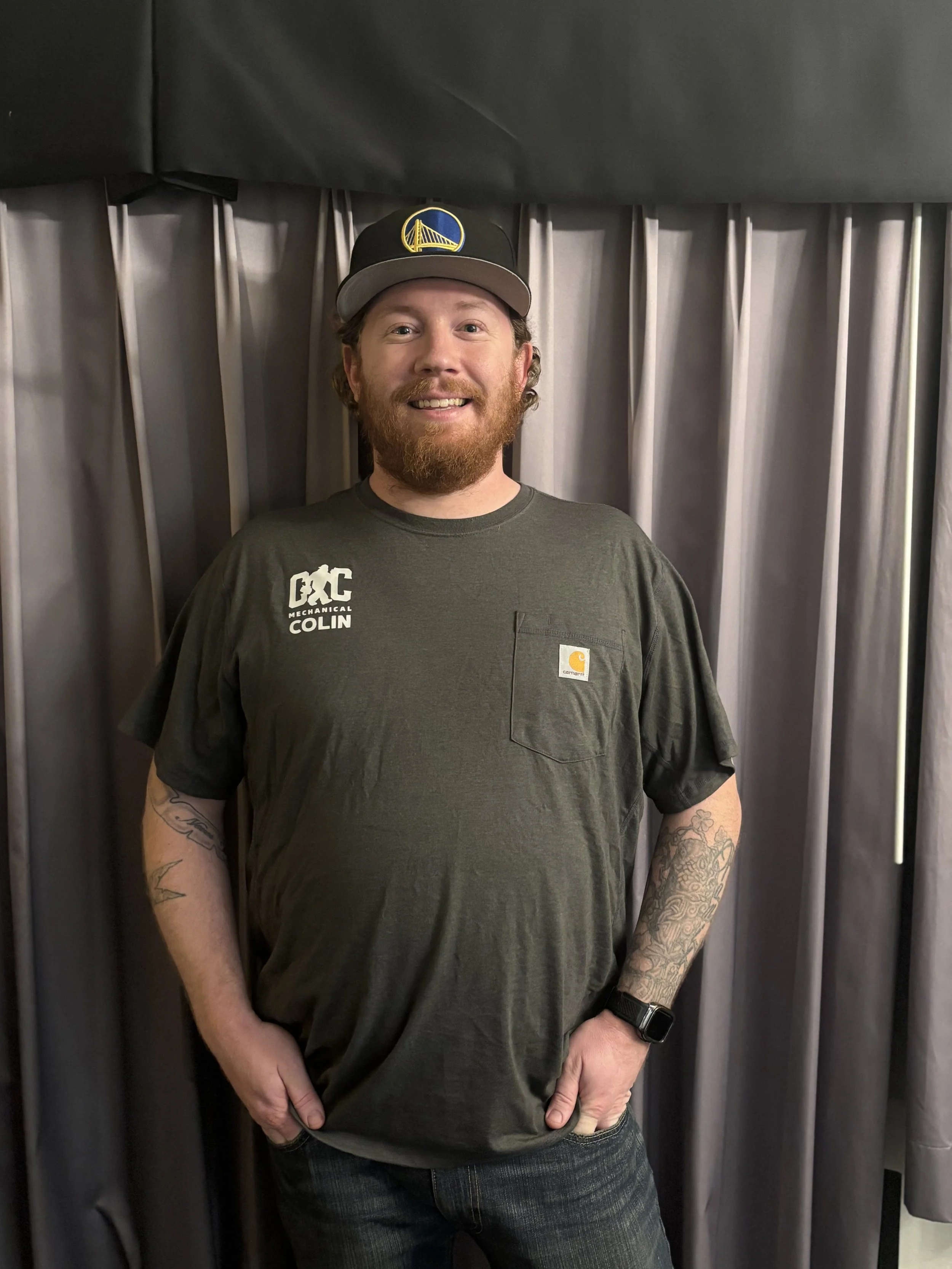 A smiling man with red hair, beard, and tattoos wearing a black hat with a Golden Gate Bridge logo, dark gray Carhartt t-shirt, and a smartwatch, posing in front of gray curtains.