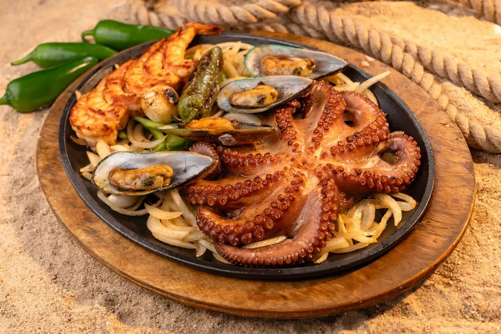 Seafood platter with octopus tentacles, shrimp, mussels, and green peppers on a bed of onions, served on a wooden plate.