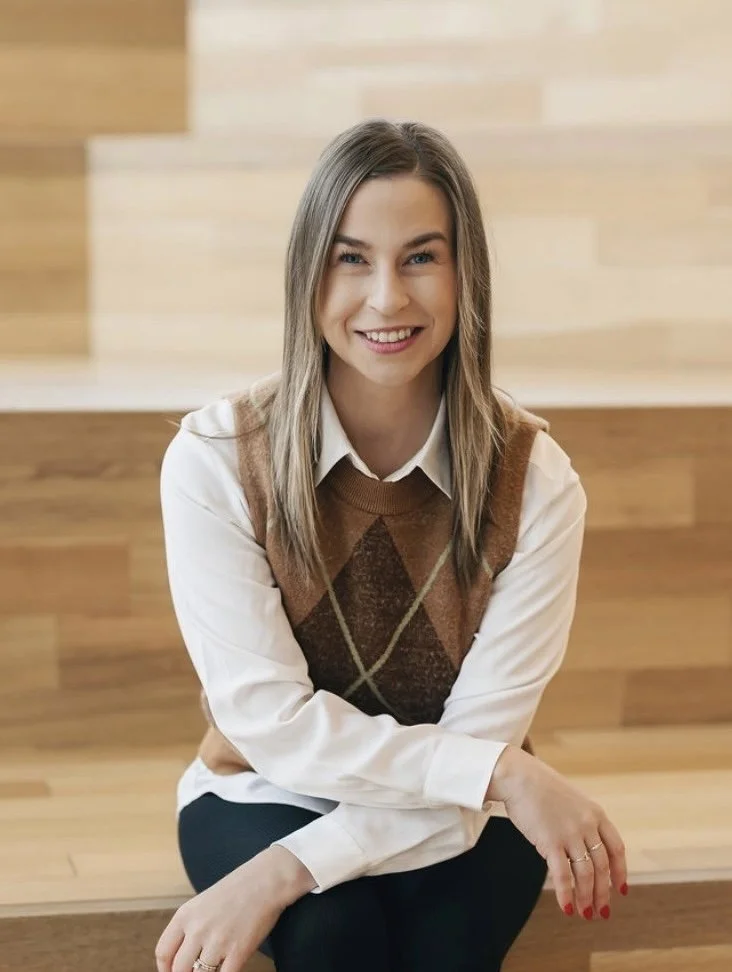 Woman in black hat and orange sweater standing in a forest, smiling under a tree with red berries.