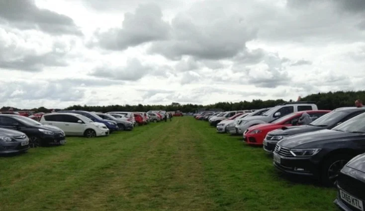 View of a grassy parking field with two rows of cars parked on either side, under a cloudy sky.