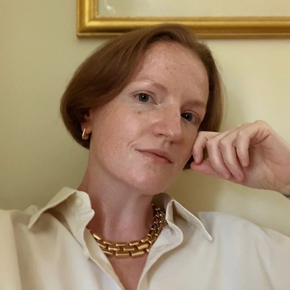 Embry has her head tilted and resting on a hand with her dark eyes looking at the camera. She wears a white collared shirt and thick gold necklace. Her read hair is cropped below the ears.