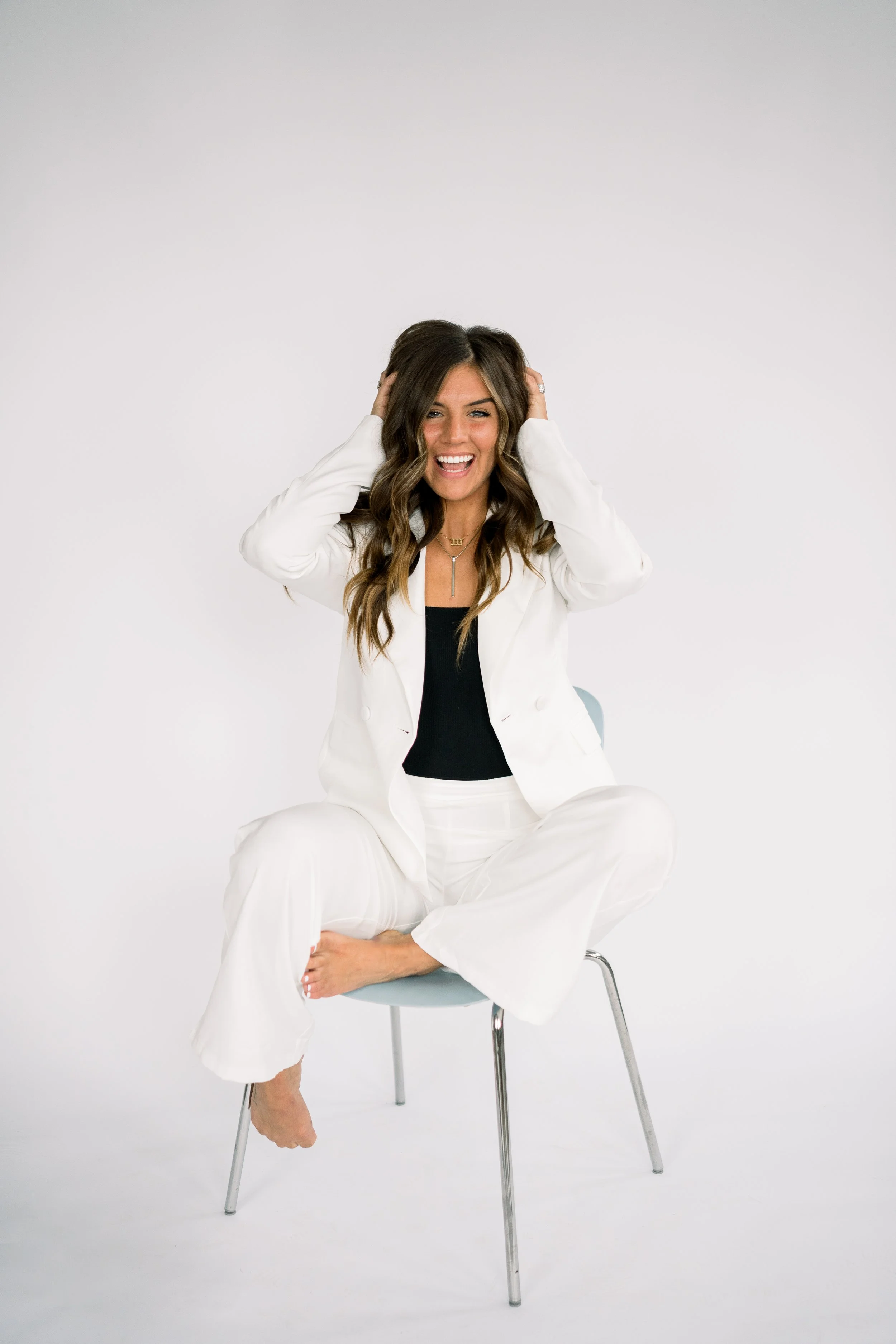 Gabby wears a white suit with a blank tank top underneath. She sits on a white chair with one foot propped on the chair under her knee with her hands in her hair. She smiles big with bright eyes against a white background.