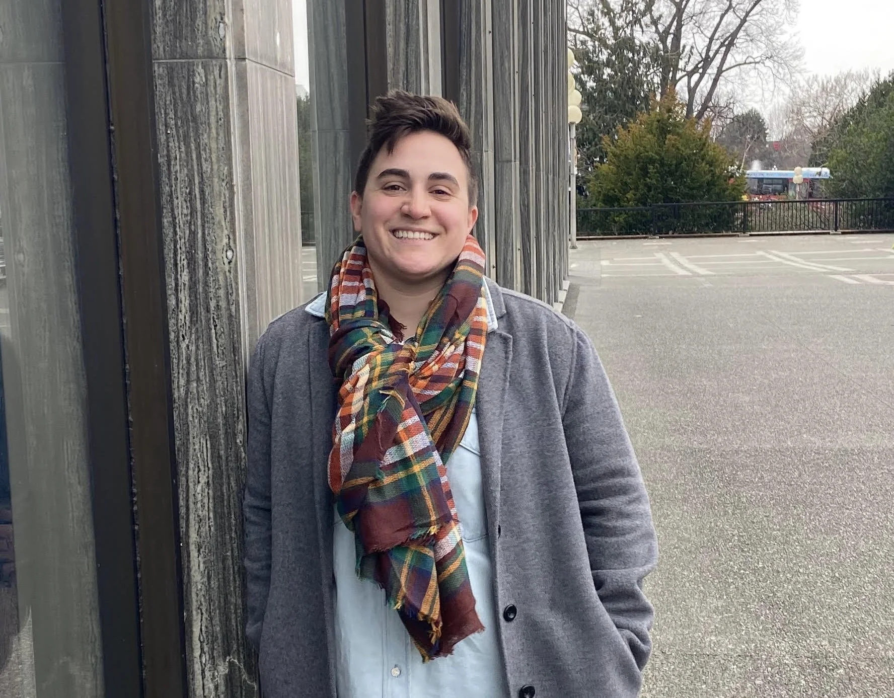Cat leans against an outdoor wall wearing a grey coat with dark buttons and a colorful plaid scarf around their neck. Cat smiles.
