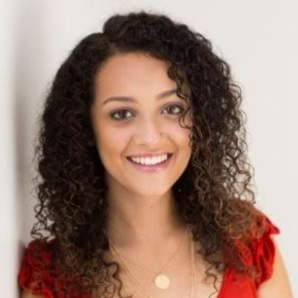 Brynn smiles and makes eye contact with the camera. Her red shirt's short sleeves peak through her curly brown hair.