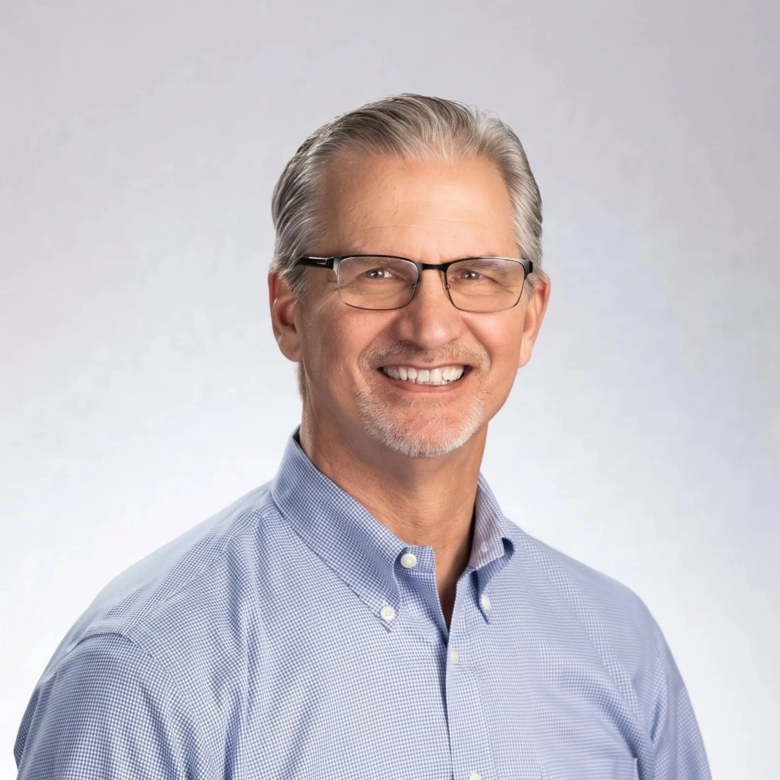 A smiling middle-aged man with gray hair, glasses, and a neatly trimmed beard, wearing a light blue dress shirt, standing against a plain light gray background.