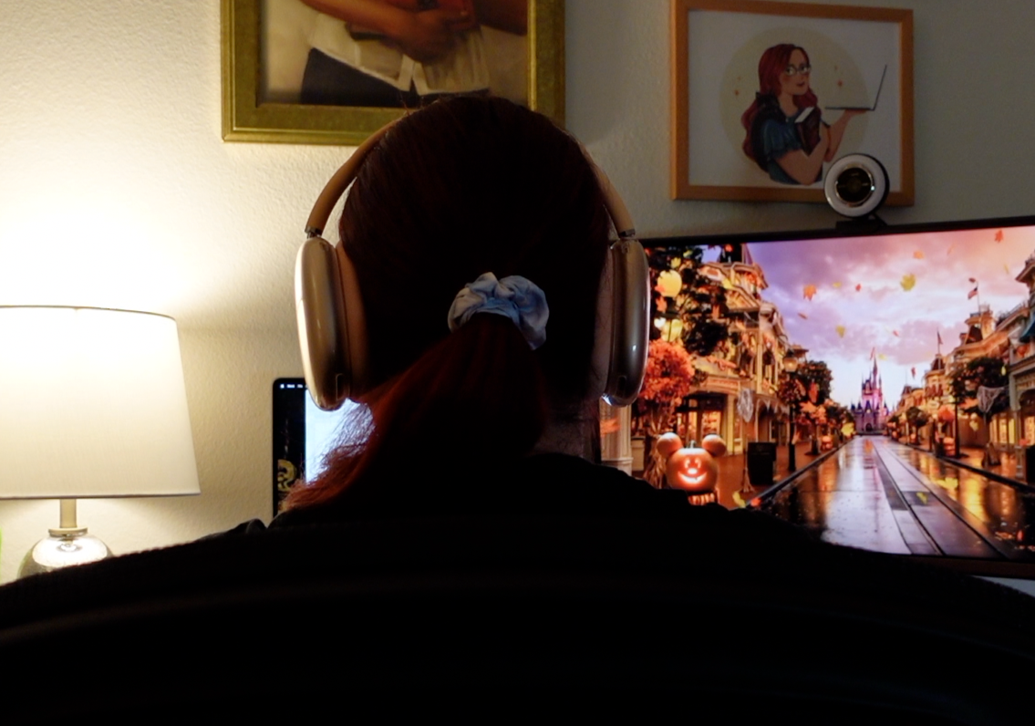 Person watching a computer screen with a Disney-themed castle scene, wearing headphones in a room with artwork and a lamp.