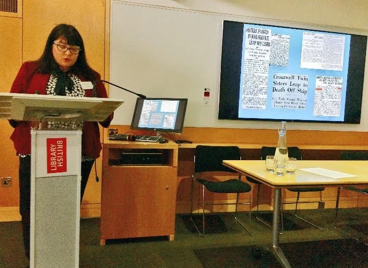 A woman standing at a podium in front of a large screen displaying a newspaper page, with a small computer on a desk nearby, in a library setting.