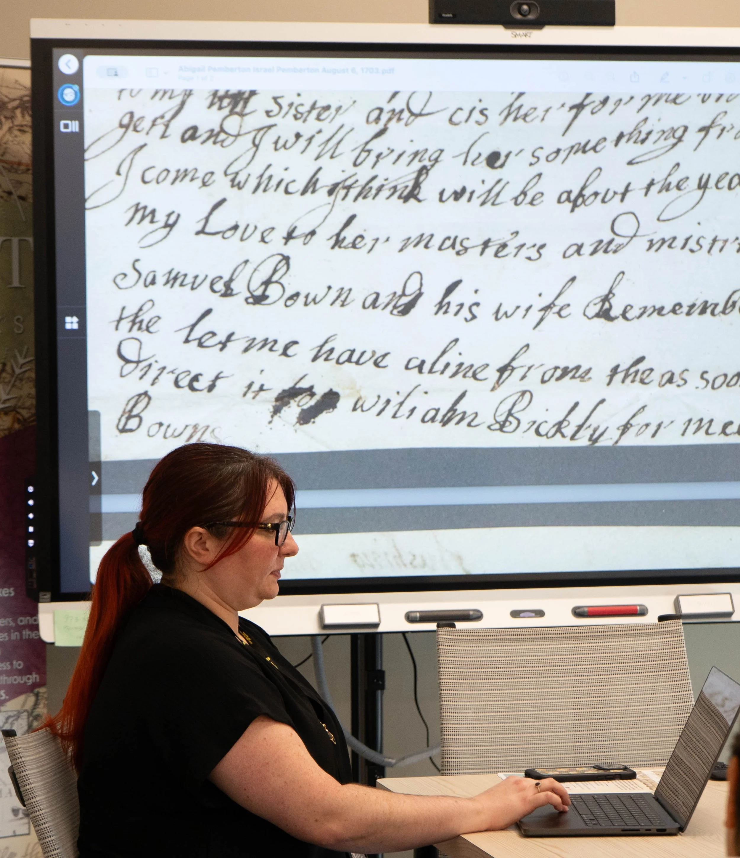 A woman with red hair tied in a ponytail, wearing glasses and a black shirt, sitting at a desk and working on a laptop in front of a large screen displaying handwritten text.