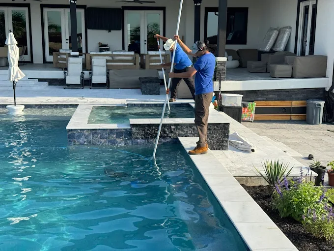 Two workers are cleaning a backyard swimming pool with a long pole, near a modern house with outdoor seating and potted plants.