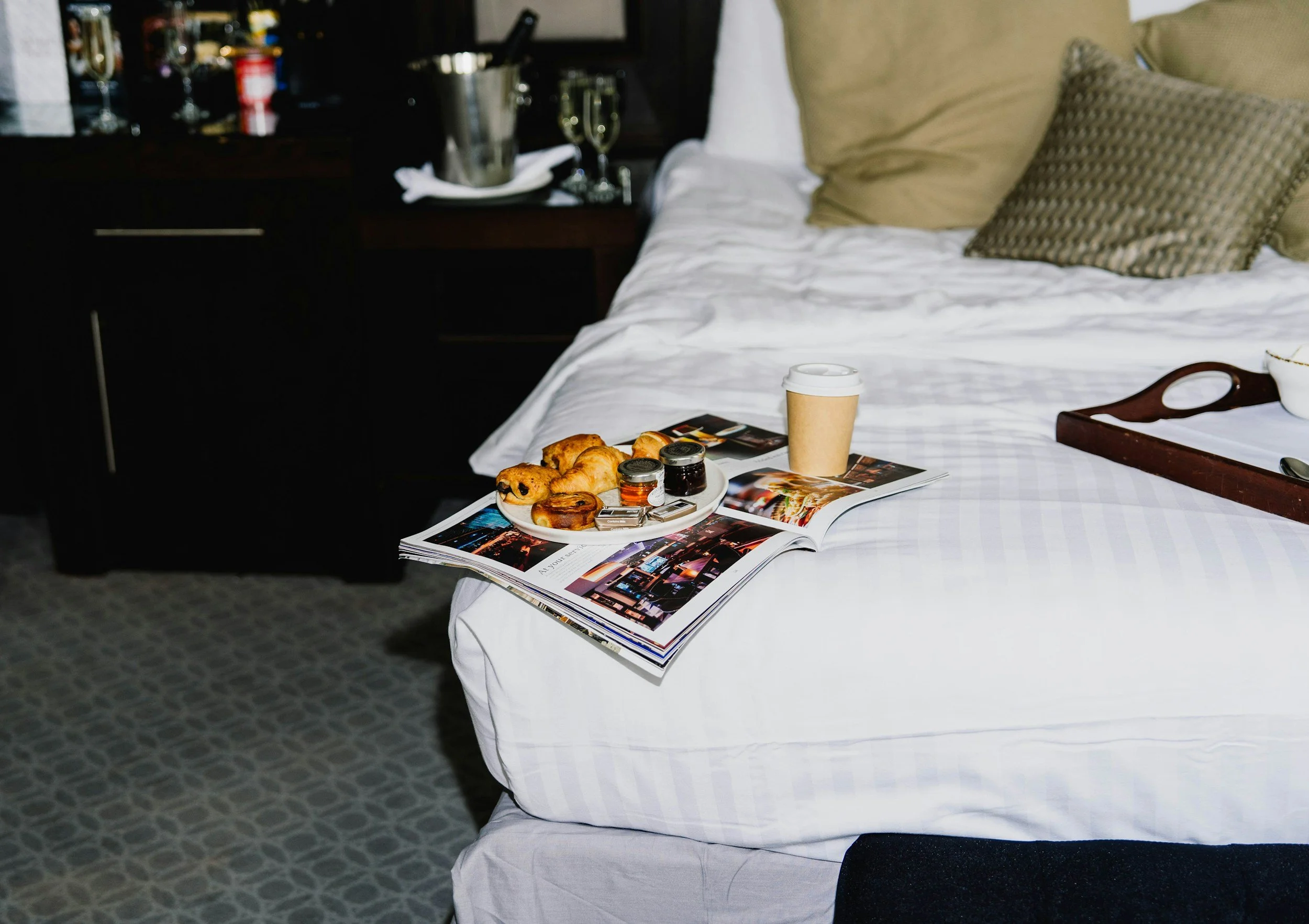 Hotel room bed with a magazine, bed tray, plate of food and a cup on top