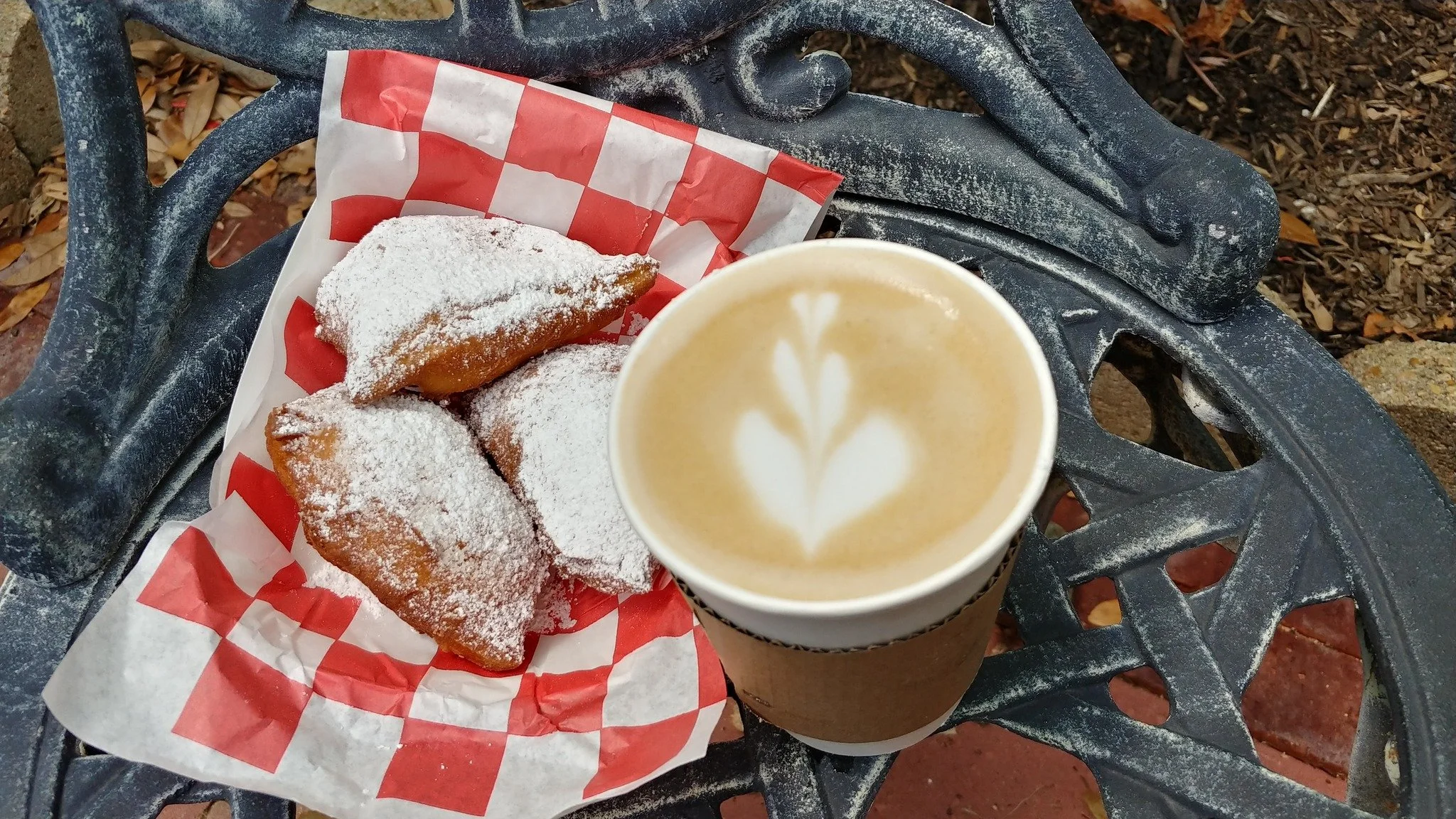 Coffee with flower design in foam next to basket of powdered sugar coated fried pies on an wrought iron chair