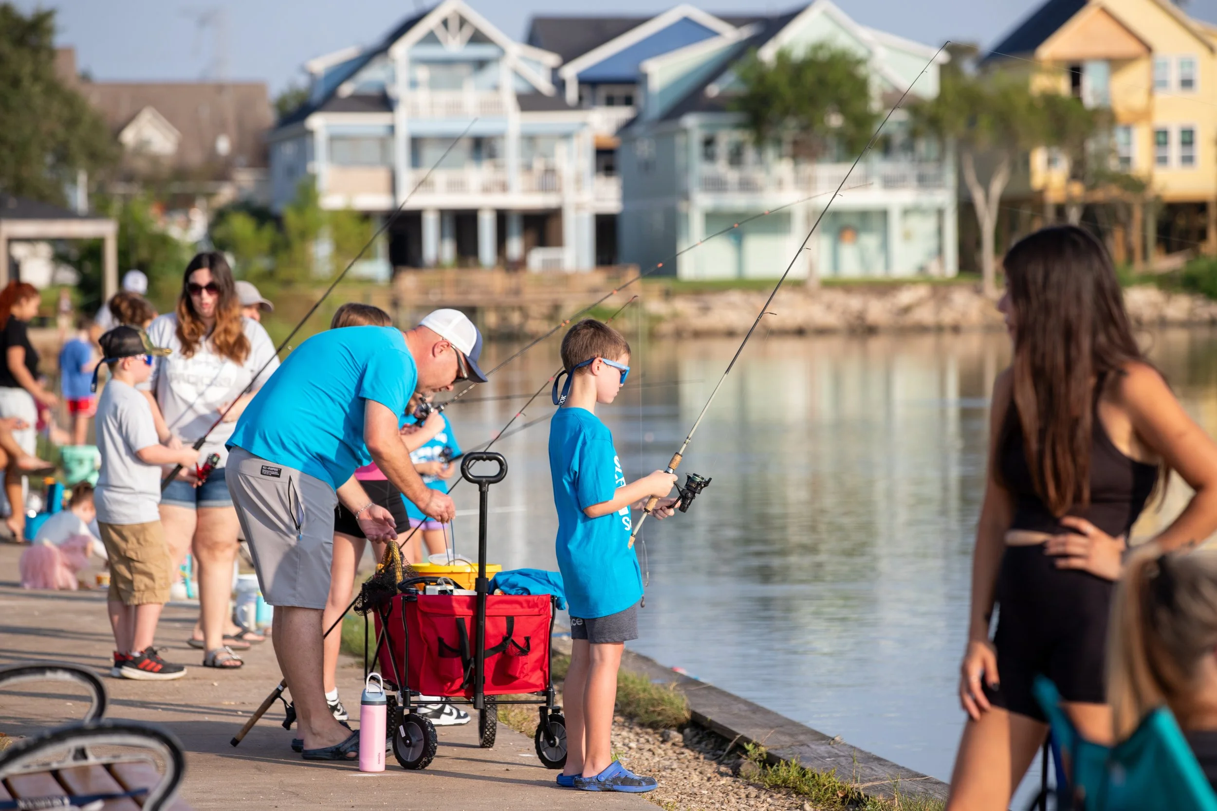 Child fishing at waterfront park w a red wagon beside him and people with fishing poles nearby