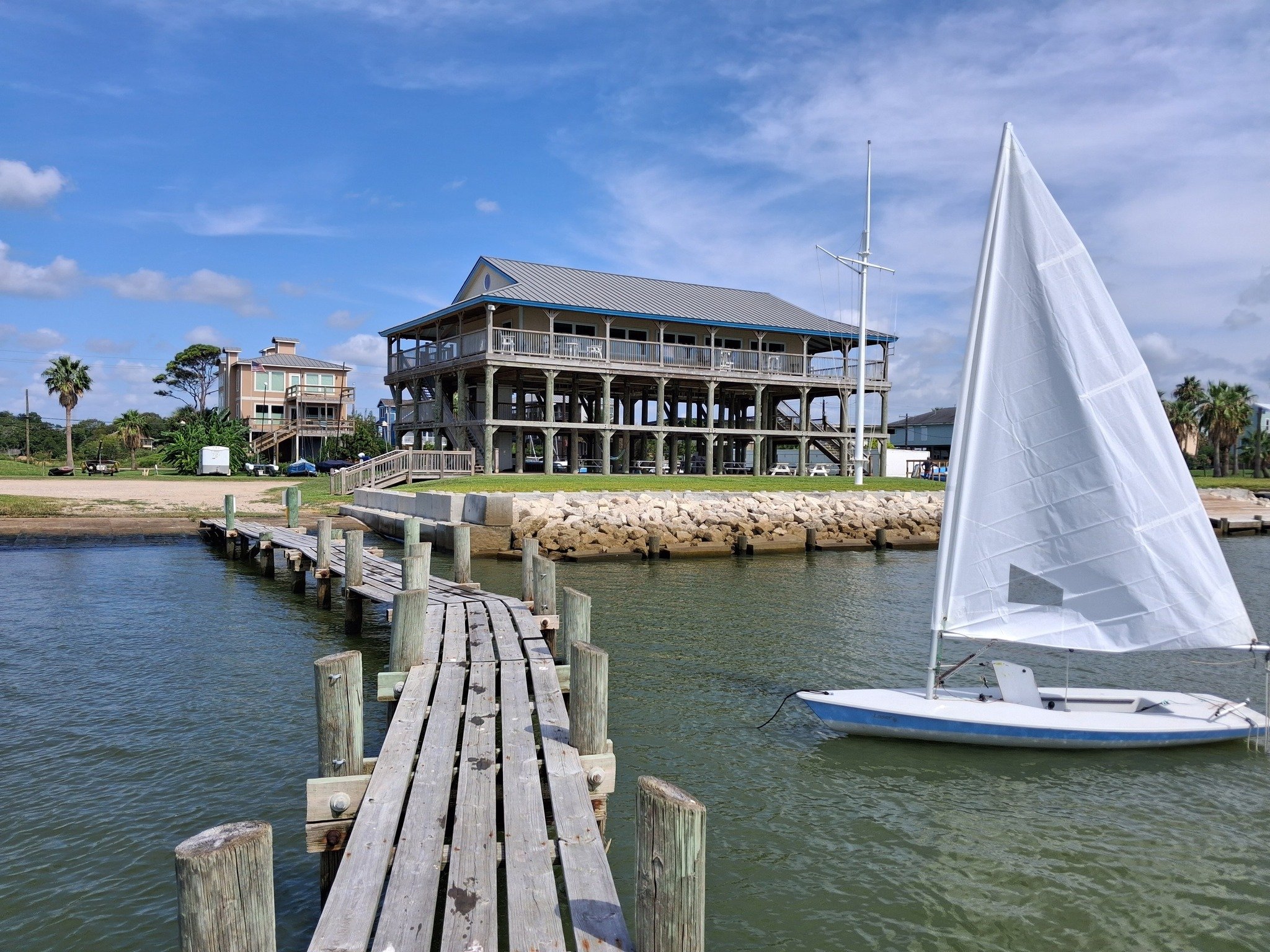 Sailboat with white sail in the water floating next to a wooden dock in front of a beach house style clubhouse