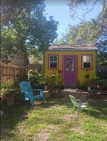 Small yellow shed with purple door in backyard with blue and green Adirondack chairs