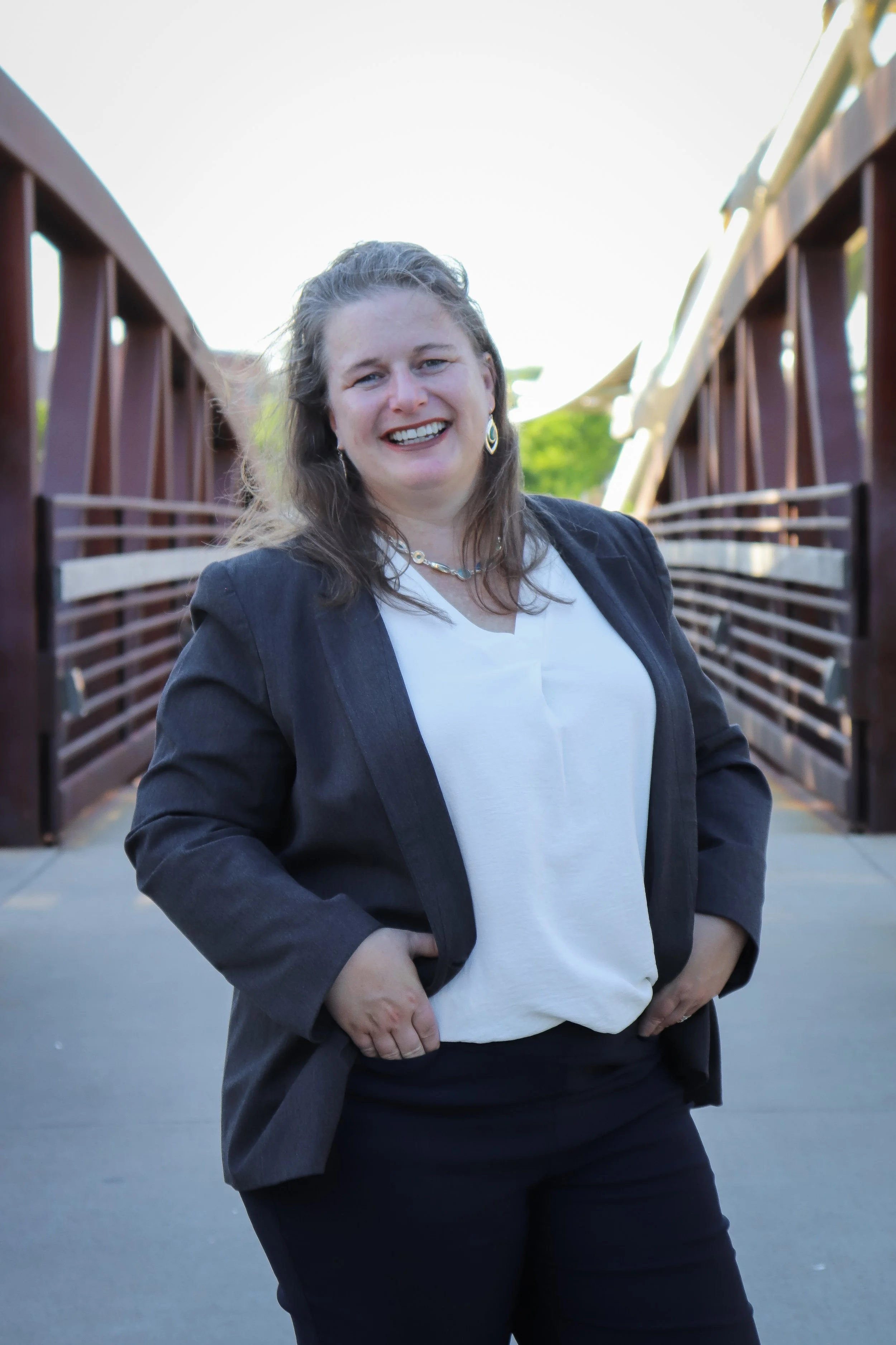Sonja Mentzer with long brown hair wearing a black blazer over a white shirt standing on a bridge with brown railings, outdoors with trees and sky in the background.