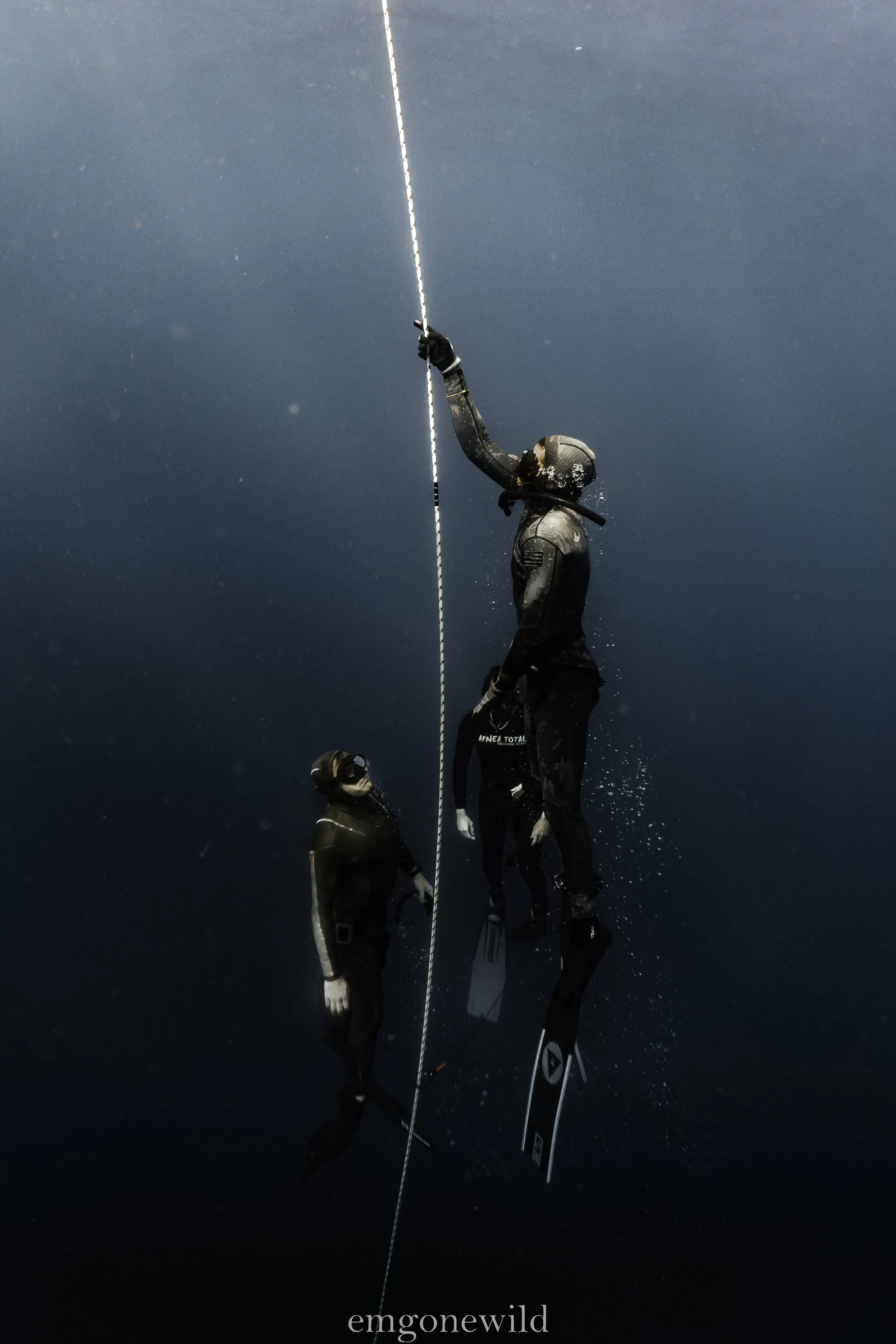 Divers underwater holding a vertical rope, wearing wetsuits and fins, with bubbles rising to the surface.