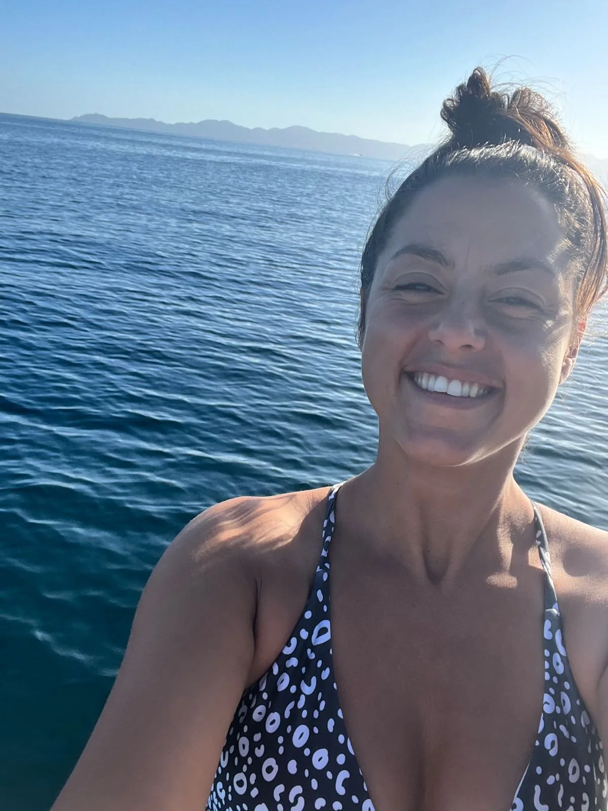 Person taking a selfie on a boat, wearing a patterned swimsuit, with the ocean and distant mountains in the background.
