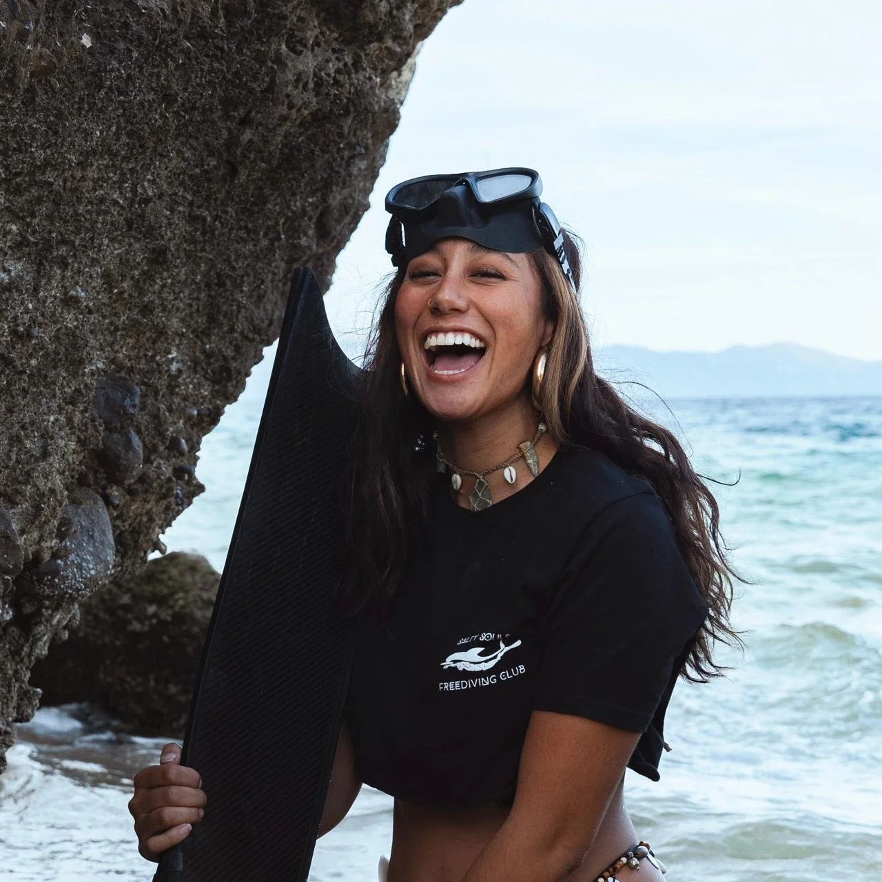 Woman in snorkel mask holding a fin, smiling on a beach near a rocky cliff.