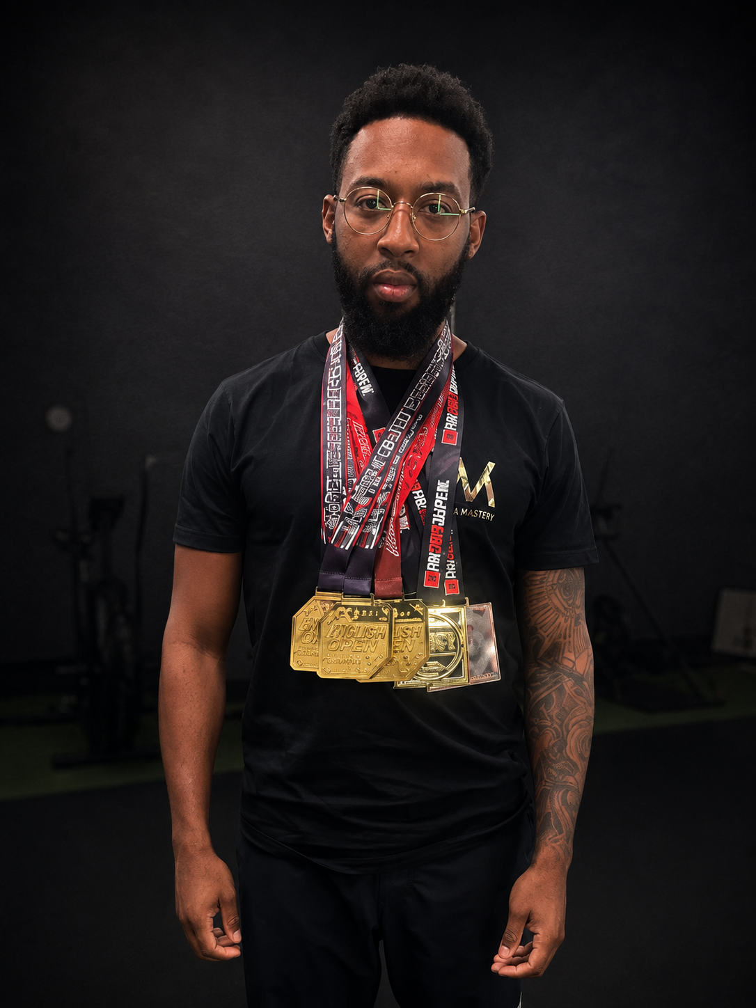 A man with glasses, a beard, and tattoos standing in a gym, wearing a black T-shirt with gold lettering and medals around his neck.