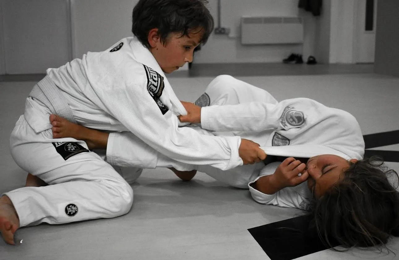 Two children practicing Brazilian Jiu-Jitsu on a gray mat; one child is on top gripping the collar of the other who is lying on the ground with eyes closed.