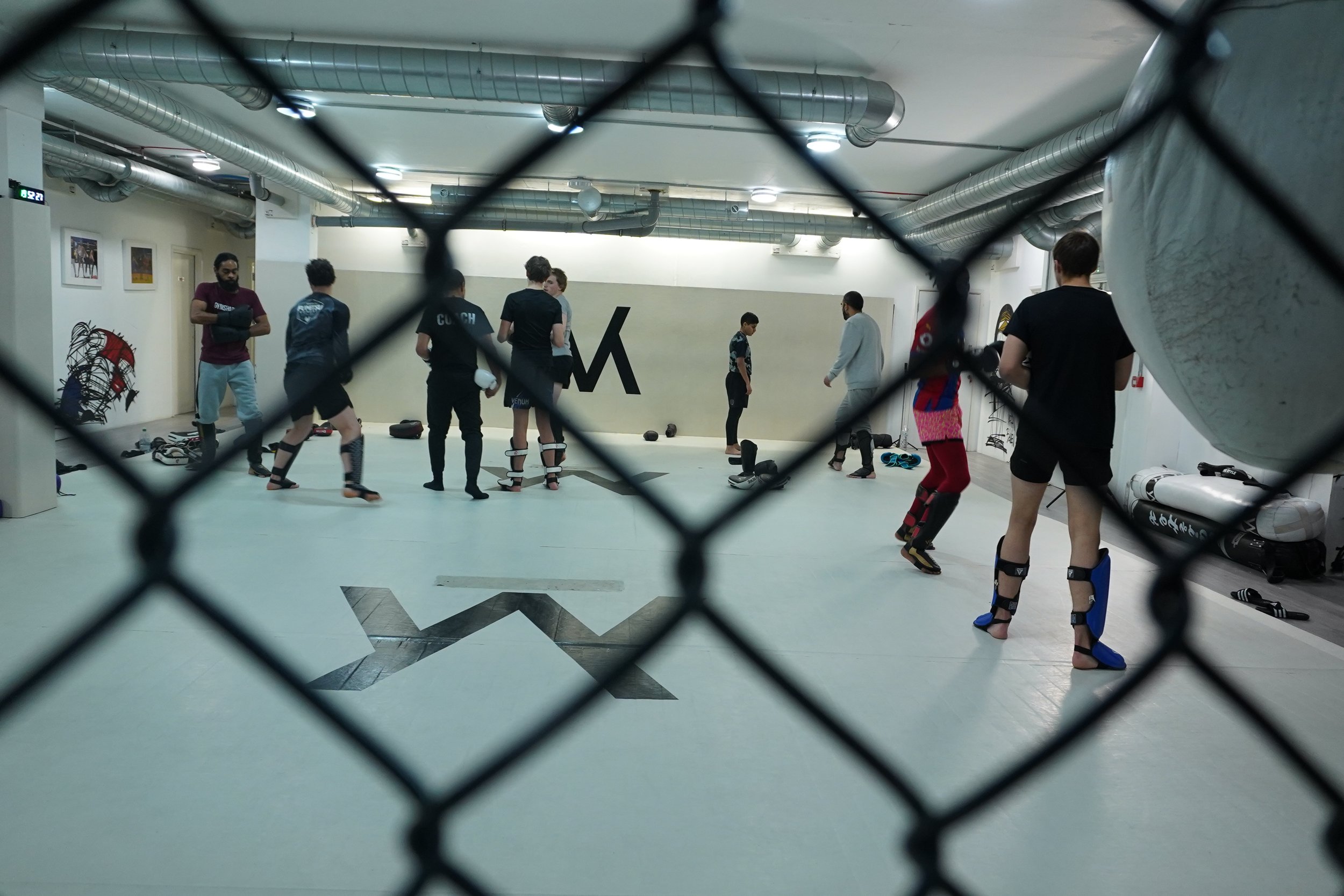 A group of people practicing martial arts or kickboxing in a gym, viewed through a chain-link fence.