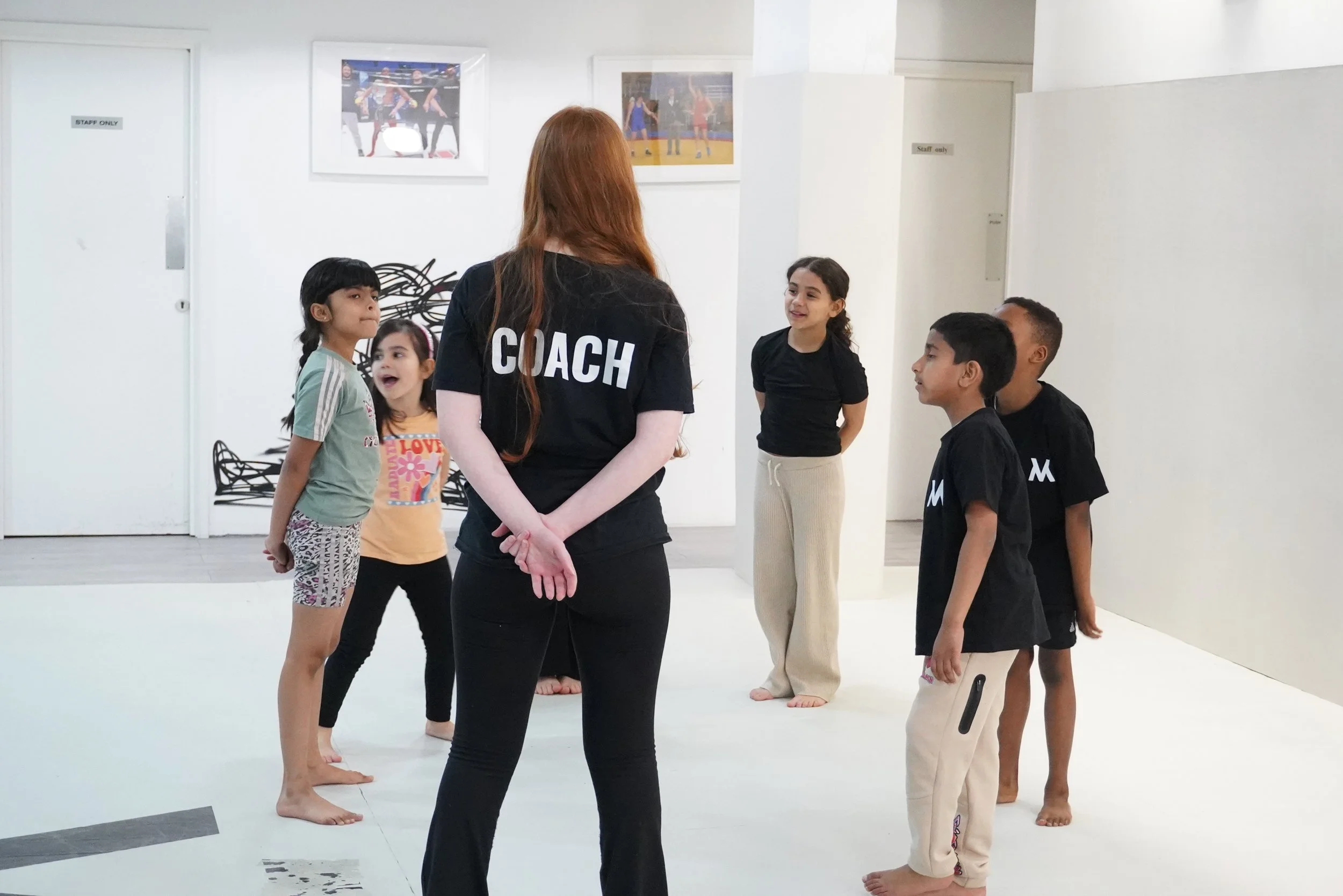 A group of children standing in a circle in a gym or studio, listening to a trainer or coach with red hair, wearing a black shirt with 'COACH' written on the back, guiding them. The children are barefoot and dressed in casual sportswear.