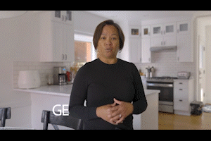 A woman standing in a modern kitchen, speaking and gesturing with her hands. The kitchen has white cabinets, a stove, and various kitchen items.