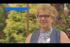 A woman with curly light hair, glasses, and a patterned blue top, standing outdoors with trees in the background.