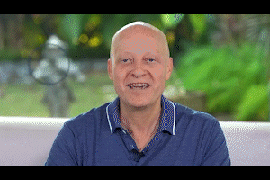 Smiling older man with short gray hair sitting indoors with a window showing greenery in the background.
