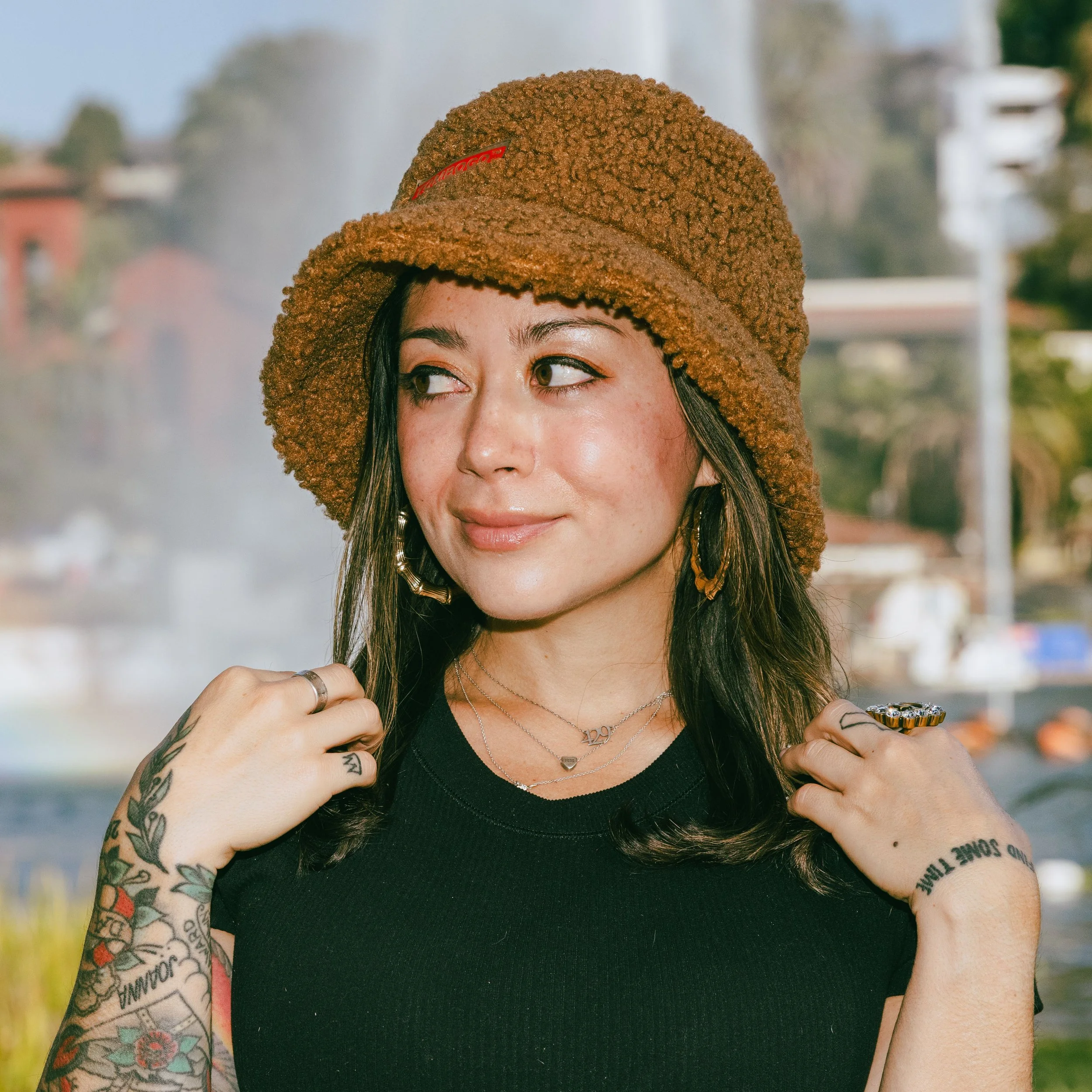 A woman with tattoos on her arms wearing a black top and a textured brown hat with a red label, walking outdoors on a sunny day.
