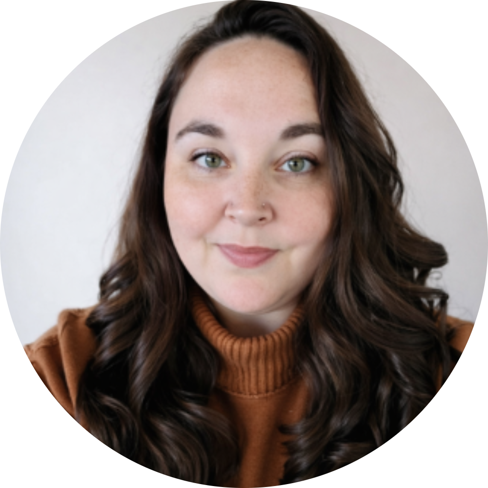 A young woman with long, curly brown hair, green eyes, and a nose piercing, wearing a brown turtleneck sweater, smiling softly at the camera.