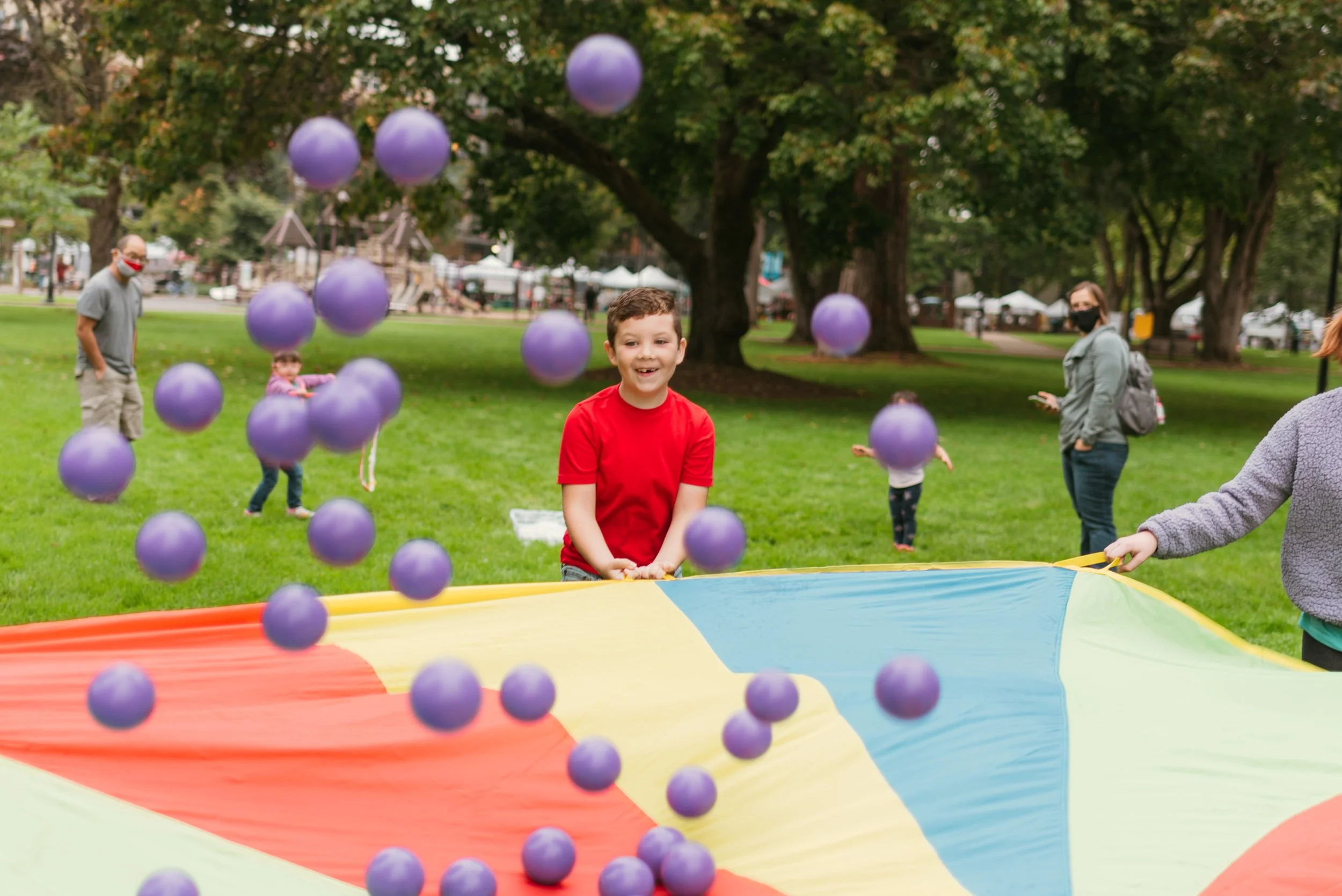Children playing with purple balls in a park with trees and people in the background.
