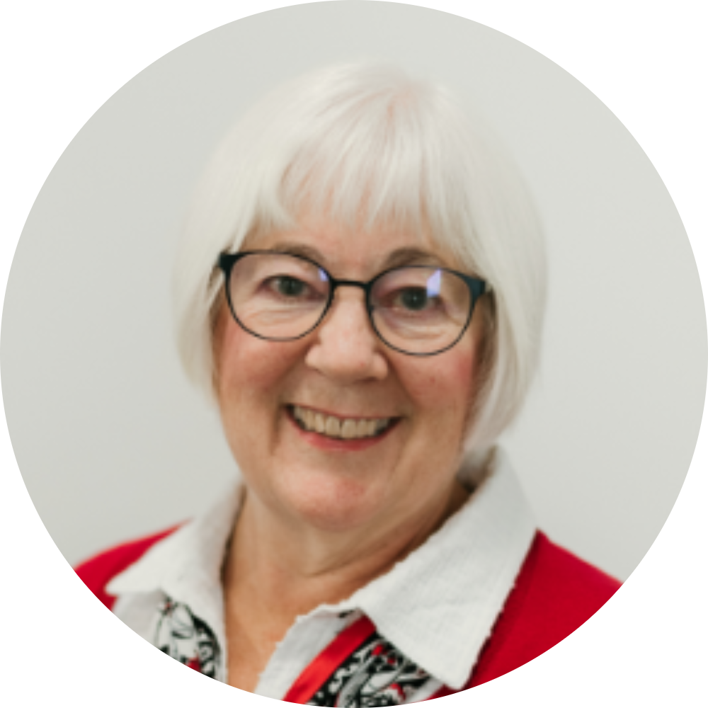 A smiling elderly woman with white hair and glasses, wearing a red blazer and a patterned shirt, in front of a plain background.