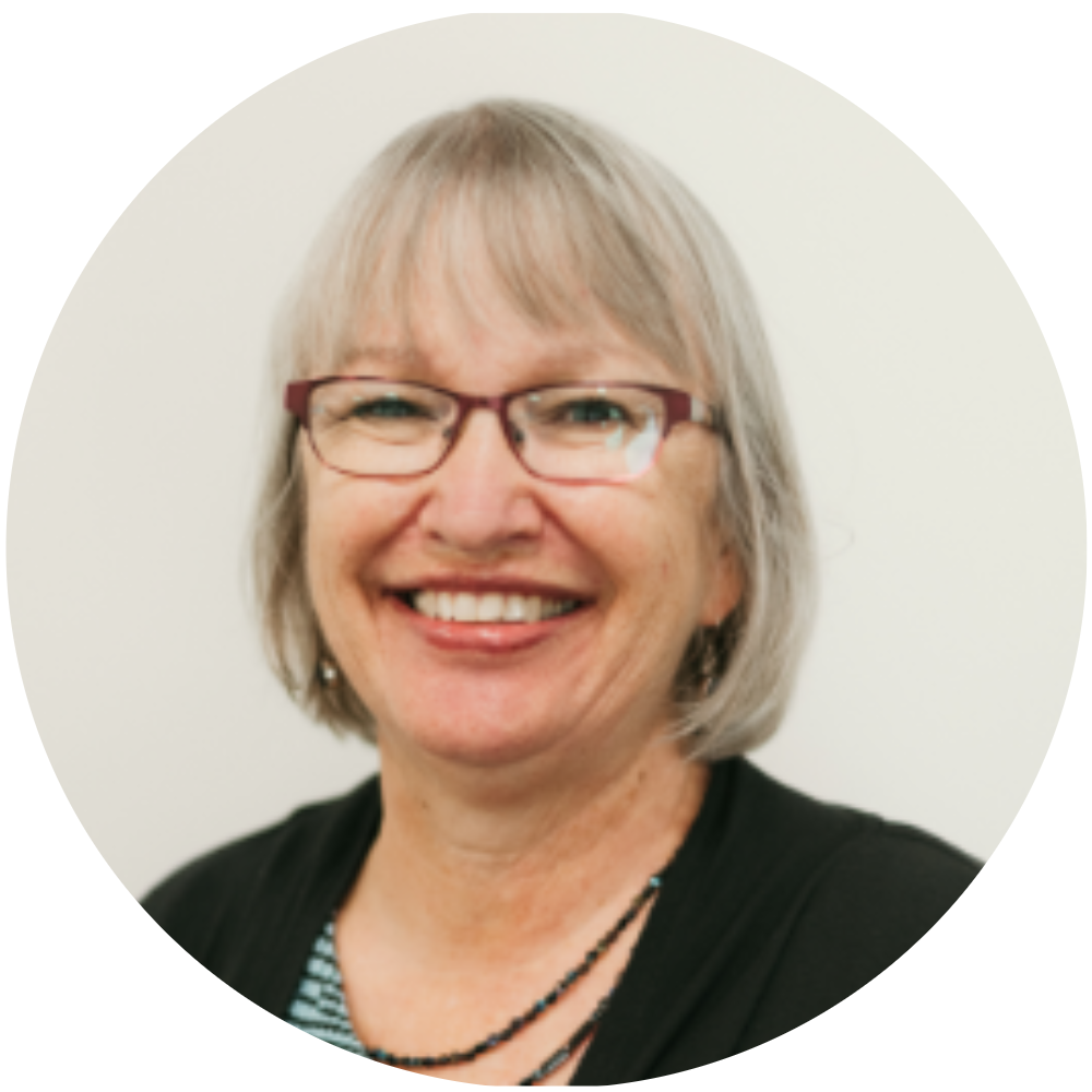 Smiling woman with short gray hair, glasses, wearing a black jacket and a necklace, standing in front of a white background with a yellow border.