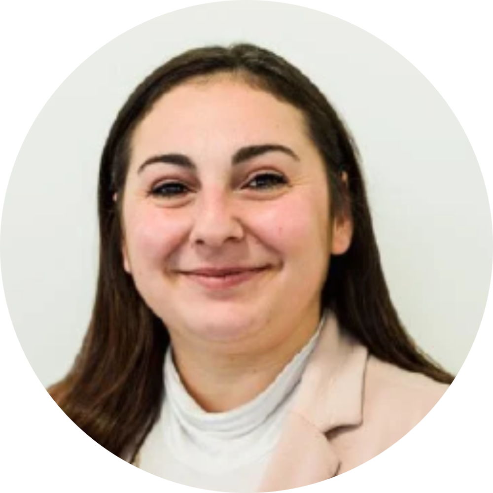 A woman with long brown hair smiling, wearing a white top and beige blazer, against a plain light background with a decorative border.