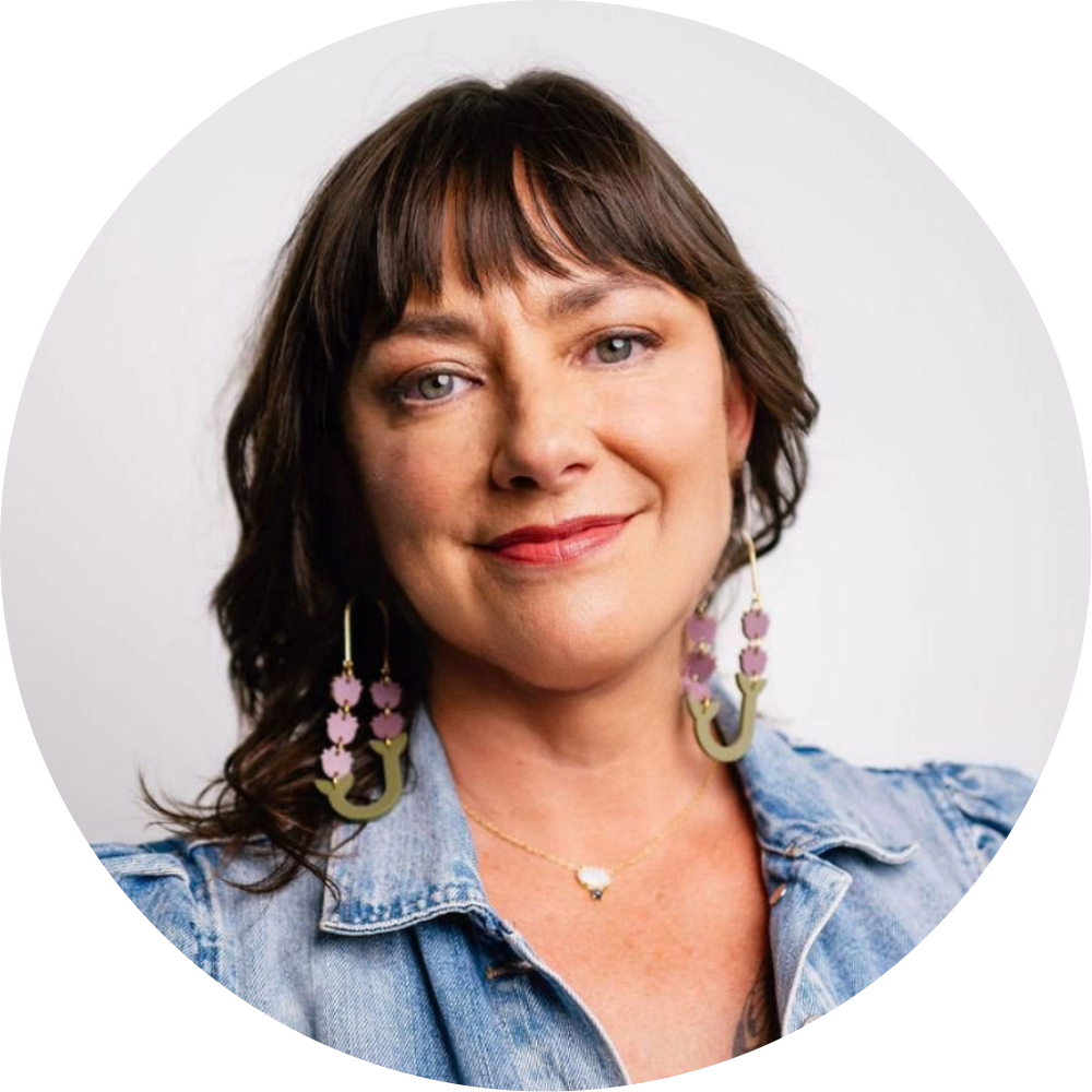 A woman with shoulder-length brown hair and bangs, wearing a denim jacket, colorful earrings, a necklace, and smiling at the camera against a plain white background.